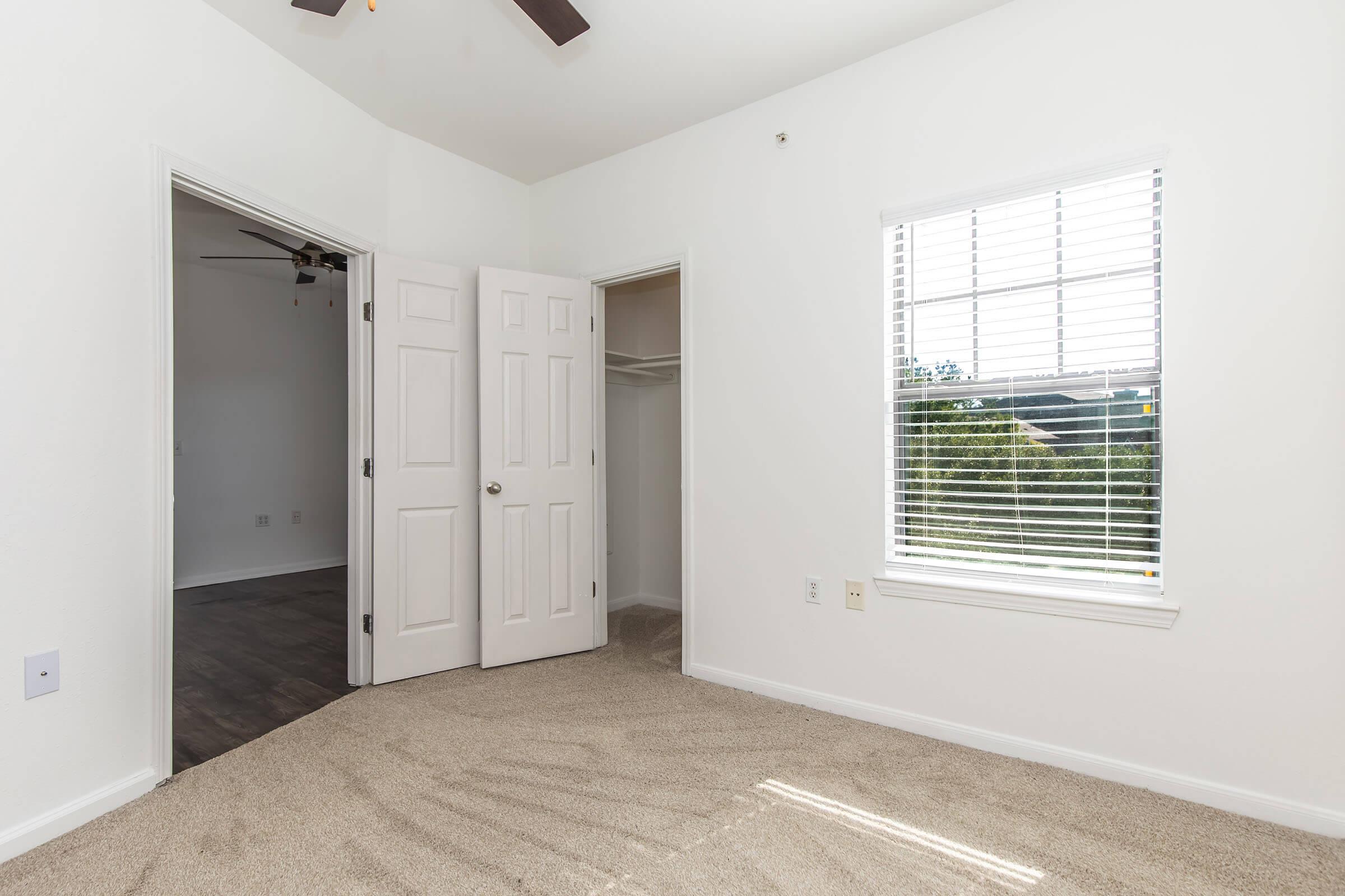 A bright, empty room featuring beige carpet, white walls, and a ceiling fan. Two doors lead to adjacent rooms, and a window with blinds allows natural light to enter. The view outside is partially visible, showcasing greenery.