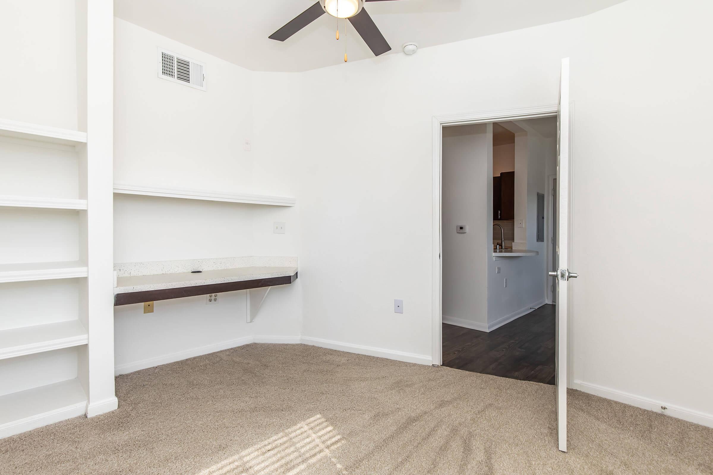 A small, empty room with beige carpet, white walls, and a ceiling fan. There is a built-in shelf and a desk space against one wall, with a door leading to another area. Light enters from a window, and the adjacent room is visible through an open door.
