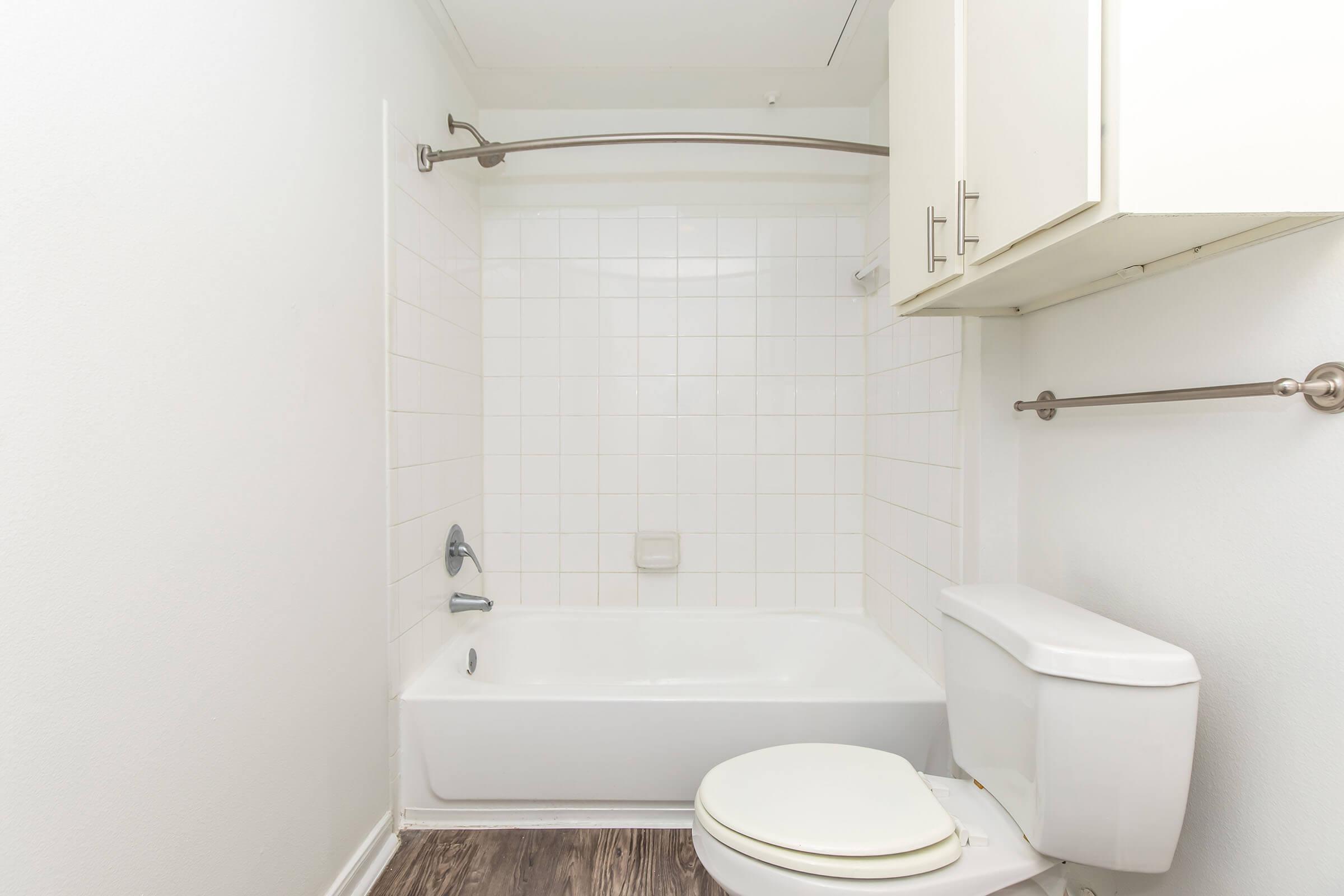 A clean, minimalistic bathroom featuring a white tiled wall surrounding a bathtub with a showerhead, a white toilet, and a small storage cabinet. The floor is a light wood laminate, and the overall color scheme is bright and neutral, creating a spacious and tidy appearance.