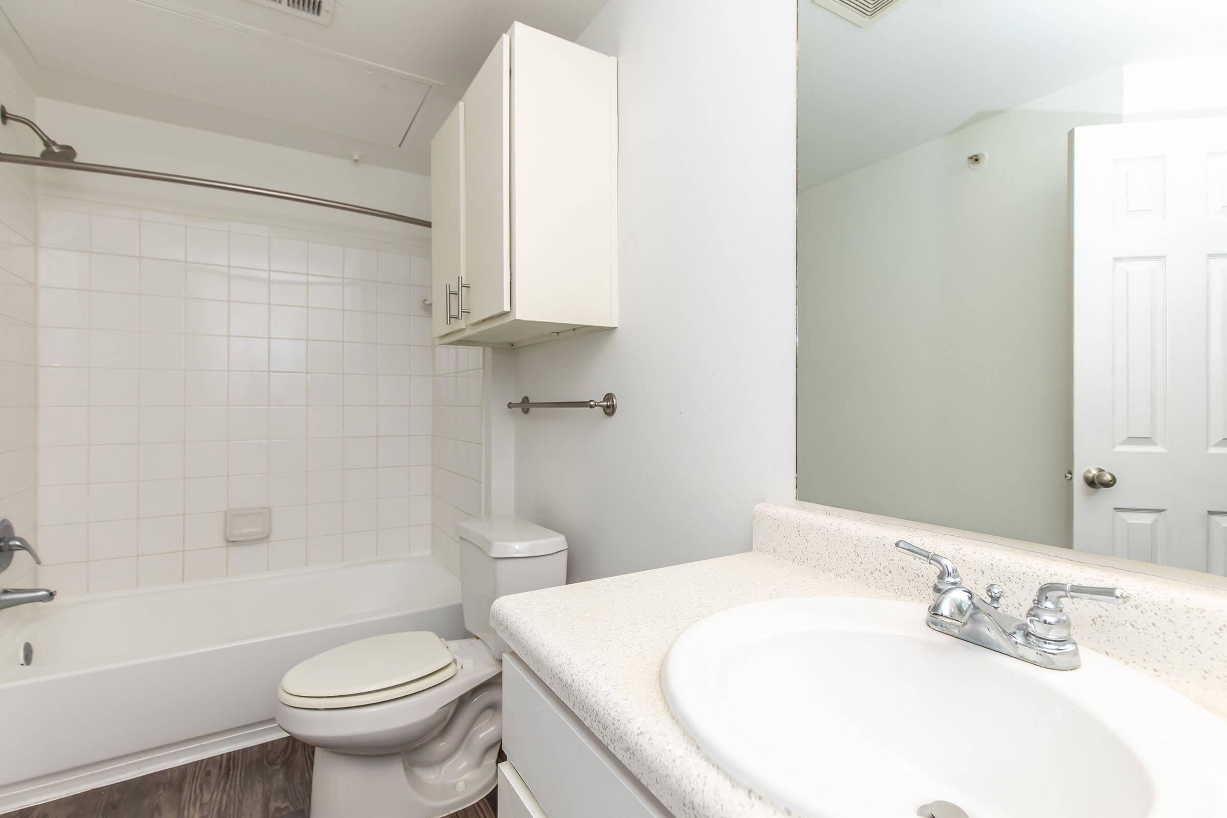 A clean bathroom featuring a white tub-shower combination, a toilet, and a sink with a countertop. The walls are light-colored, and there is a mirror visible along with a cabinet above the sink. The overall appearance is tidy and modern, with a neutral color scheme.