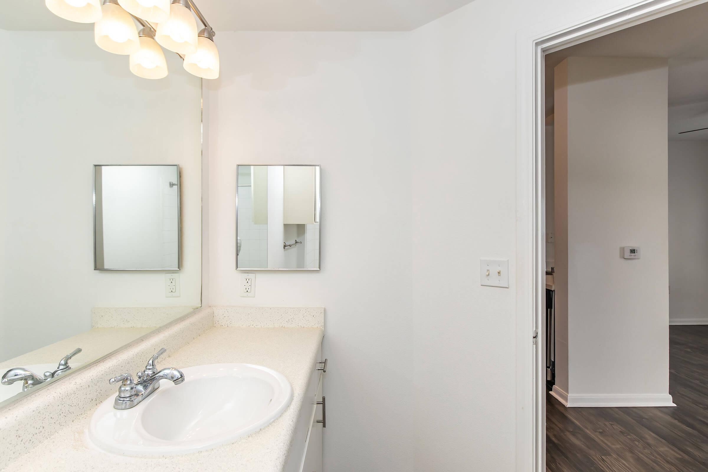 A clean bathroom featuring a white countertop with a sink, a wall-mounted mirror, and a light fixture with multiple bulbs. The walls are painted in a light shade, and there is a doorway leading to another room in the background, highlighted by wooden flooring.