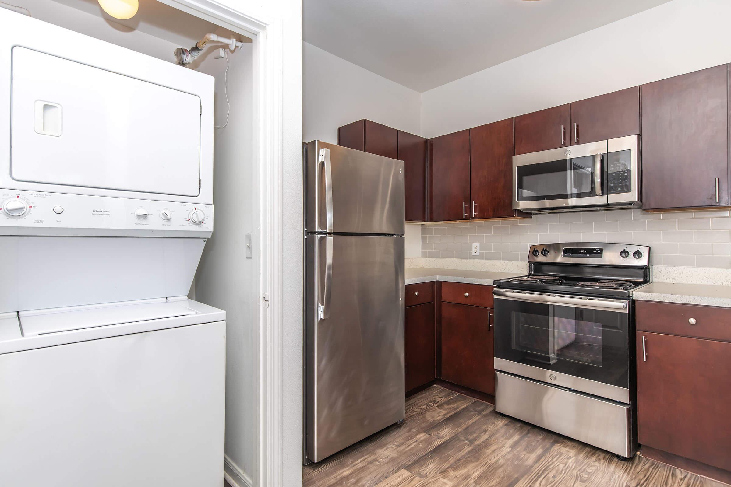 A modern kitchen featuring dark wood cabinets, a stainless steel refrigerator, an oven with a stove top, and a light-colored countertop. To the left, there is a stacked washer and dryer unit. The flooring is a wood-like laminate, and the space is well-lit with natural light.