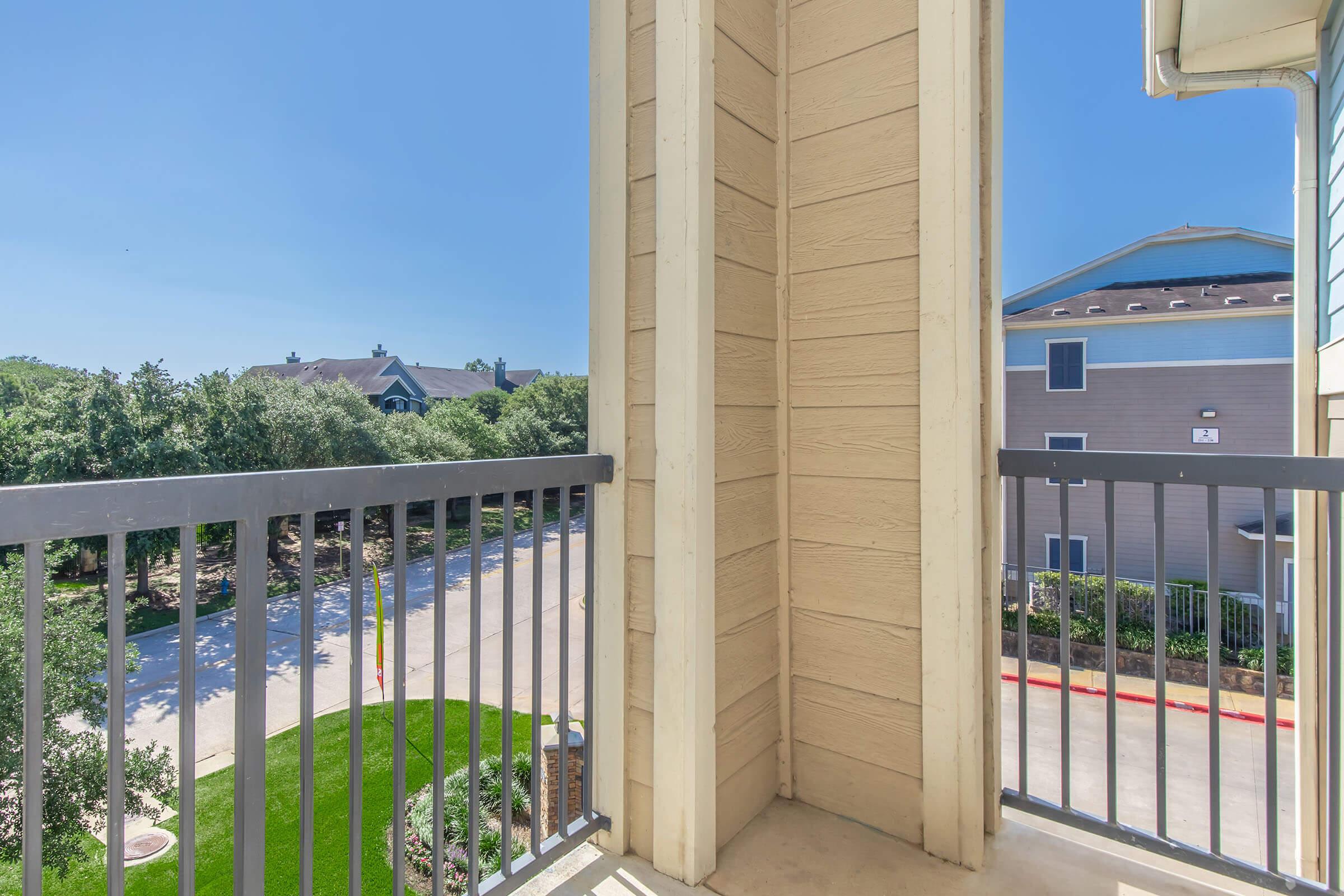A view from a balcony overlooking a green lawn and nearby buildings under a clear blue sky. The balcony features a railing and a corner that frames the scene, creating a sense of openness and tranquility in a residential area.