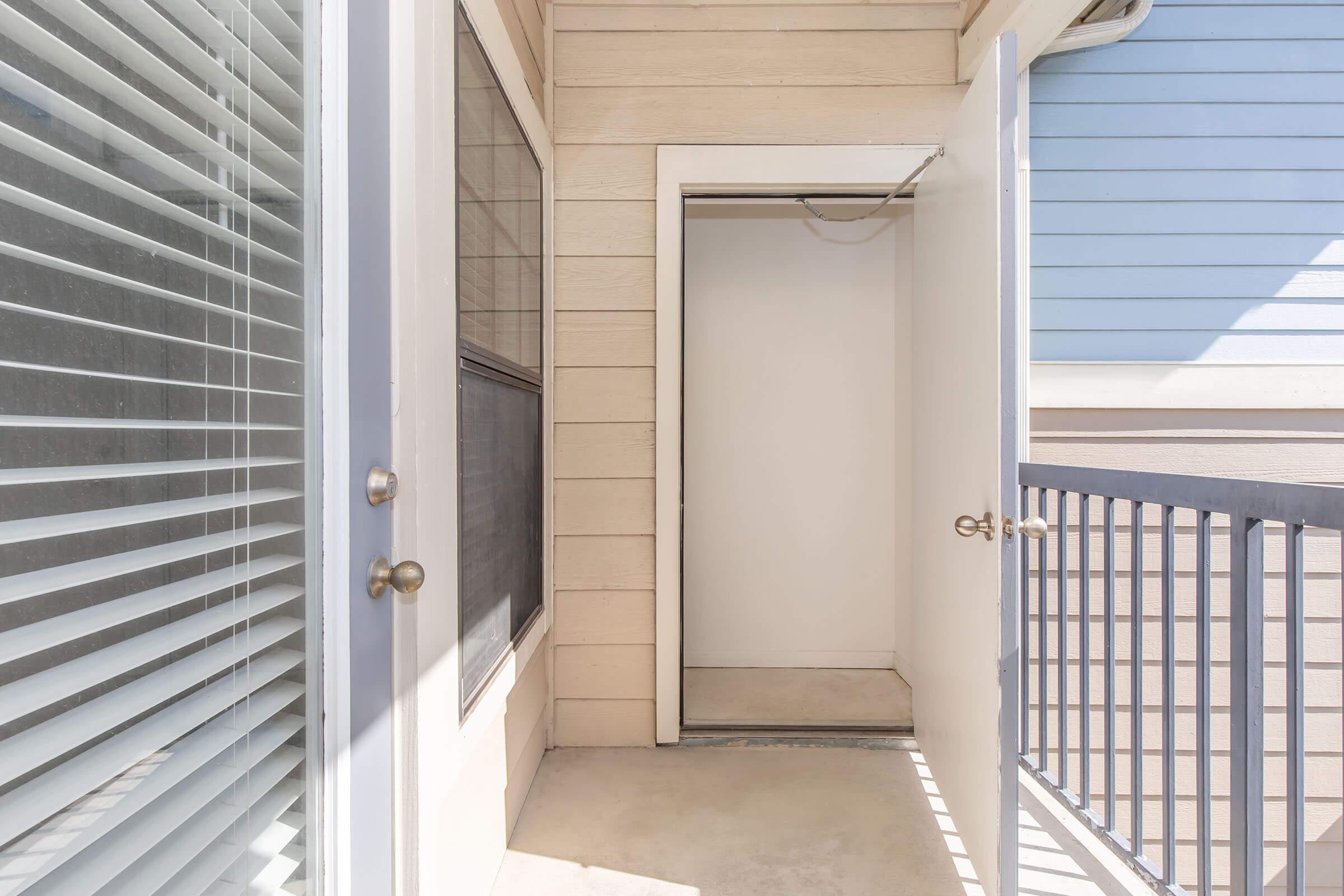 A small balcony area with a door leading to a storage closet. The wall is light-colored with horizontal siding, and there are vertical blinds on the left side. The entrance is empty and bright, with a railing visible in the foreground, creating an open and airy feel to the space.
