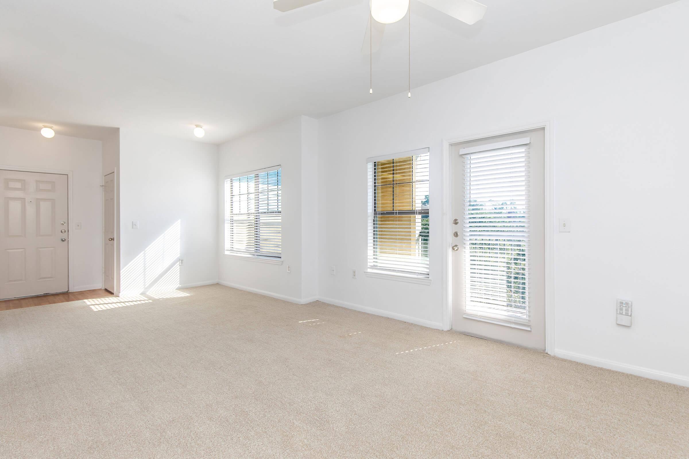 A bright, empty living room featuring light-colored walls and carpet. Windows allow natural light to enter, and a ceiling fan is visible. A door leads to a small balcony or patio area. The space is clean and spacious, ideal for furnishing and decoration.
