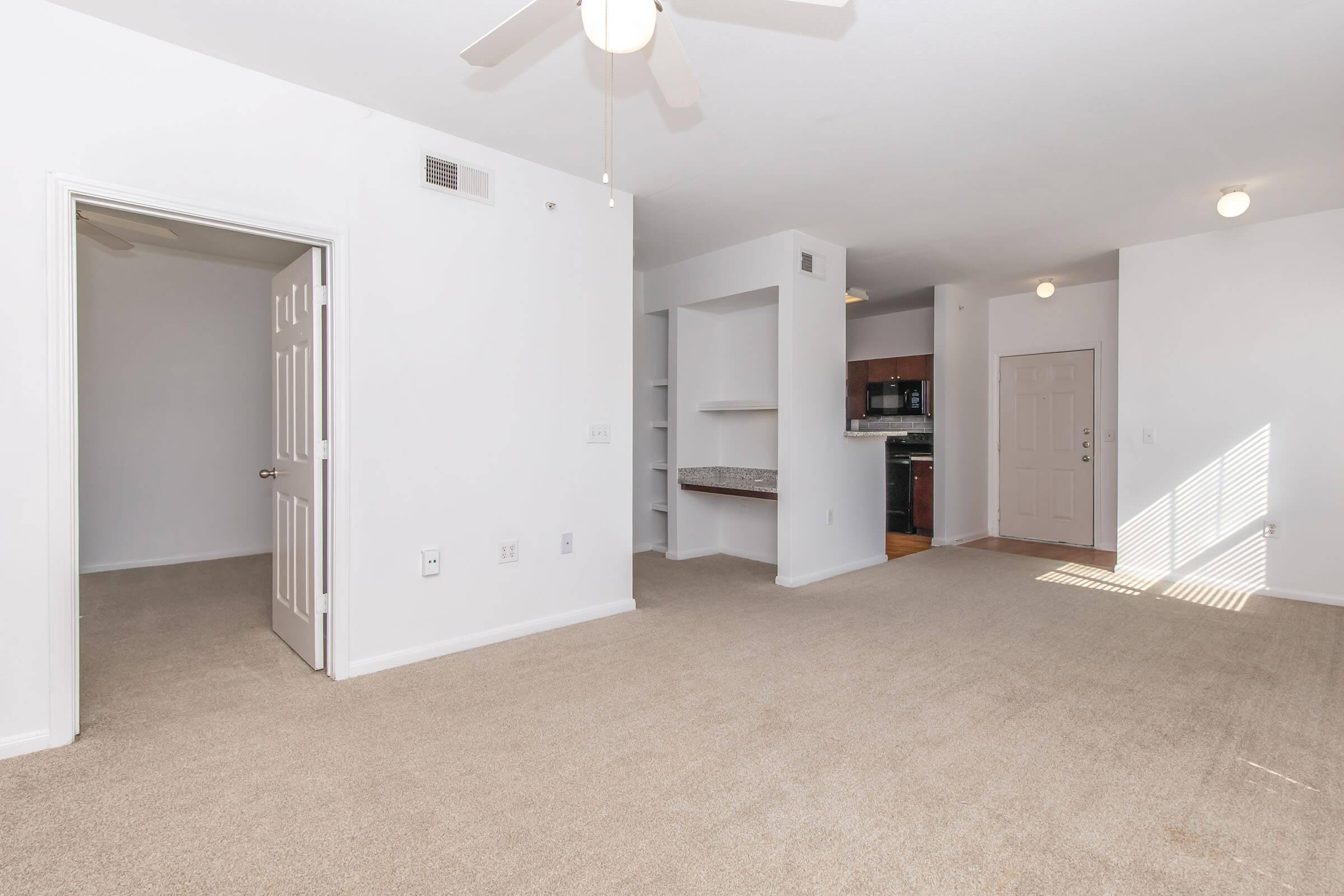 A spacious, well-lit living area with light beige carpet and neutral-colored walls. There are two doorways visible, one leading to a room and the other to a kitchen area with dark cabinetry. Natural light comes through a window, and a ceiling fan is present in the corner.
