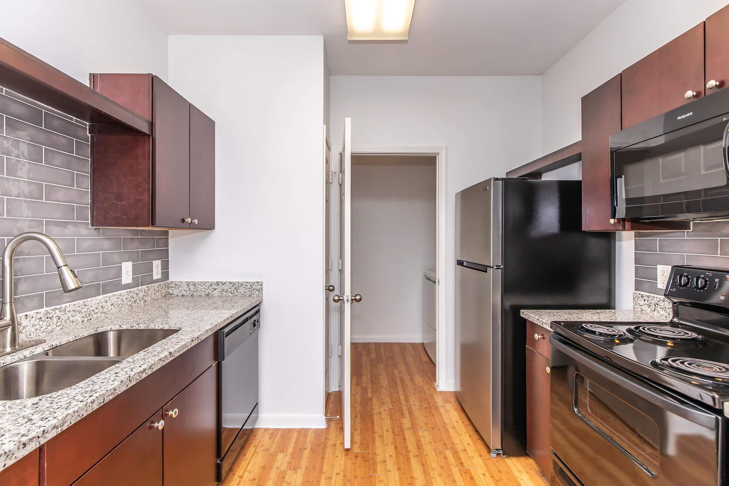 A modern kitchen featuring dark wood cabinetry, a granite countertop, and stainless steel appliances. The space includes a sink, dishwasher, electric stove, and refrigerator. A doorway leads to an adjacent room, with bamboo flooring and neutral-colored walls enhancing the contemporary feel of the kitchen.