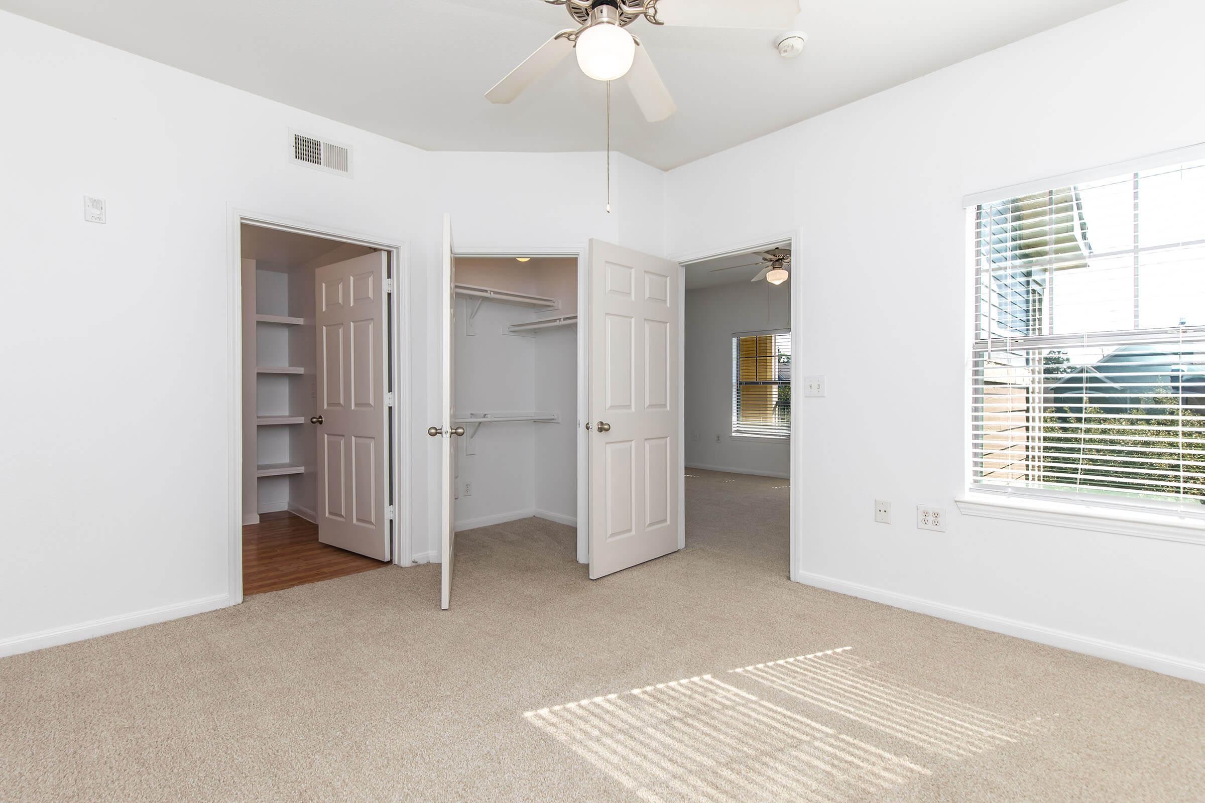 A bright and spacious bedroom featuring beige carpet, a ceiling fan, and two open doors leading to a closet and another room. Large windows allow natural light to fill the space, providing a cozy and inviting atmosphere.