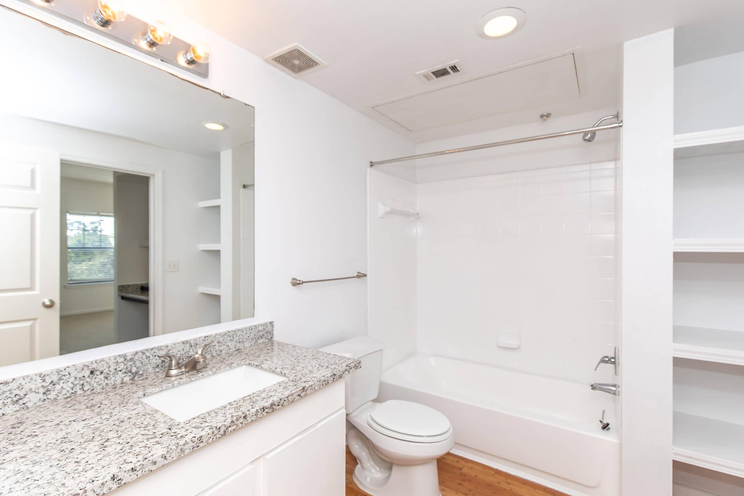A bright, modern bathroom featuring a white bathtub with a shower, a granite countertop sink, a large mirror with overhead lighting, and light-colored walls. There are built-in shelves on one side and a doorway leading to another room in the background. Natural light is coming through a window.
