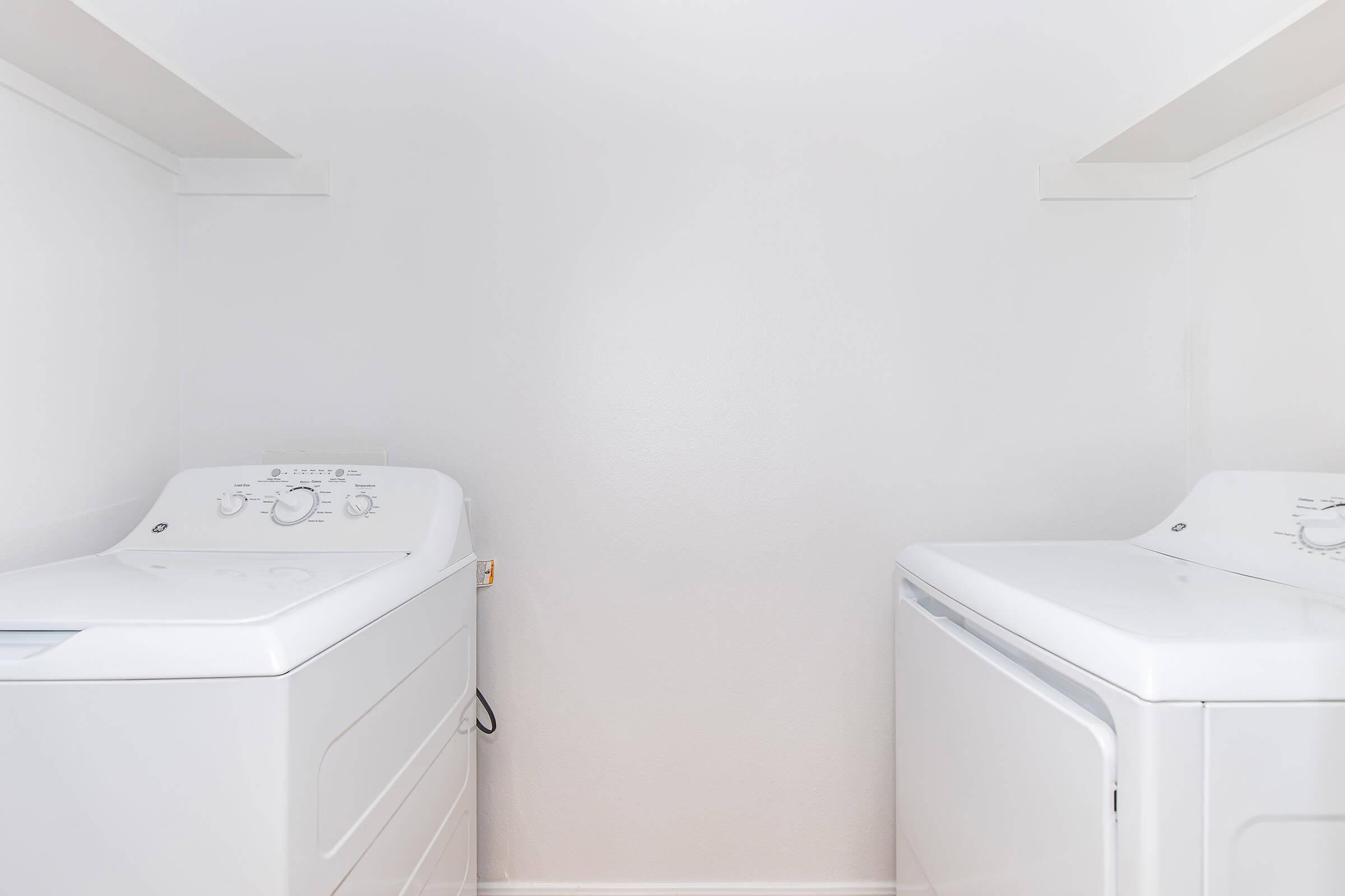 Two white laundry appliances are positioned side by side in a bright, minimalistic laundry room. The left side features a washing machine with control dials, while the right side displays a dryer. The walls are painted in a light shade, emphasizing a clean and organized space with ample room between the appliances.