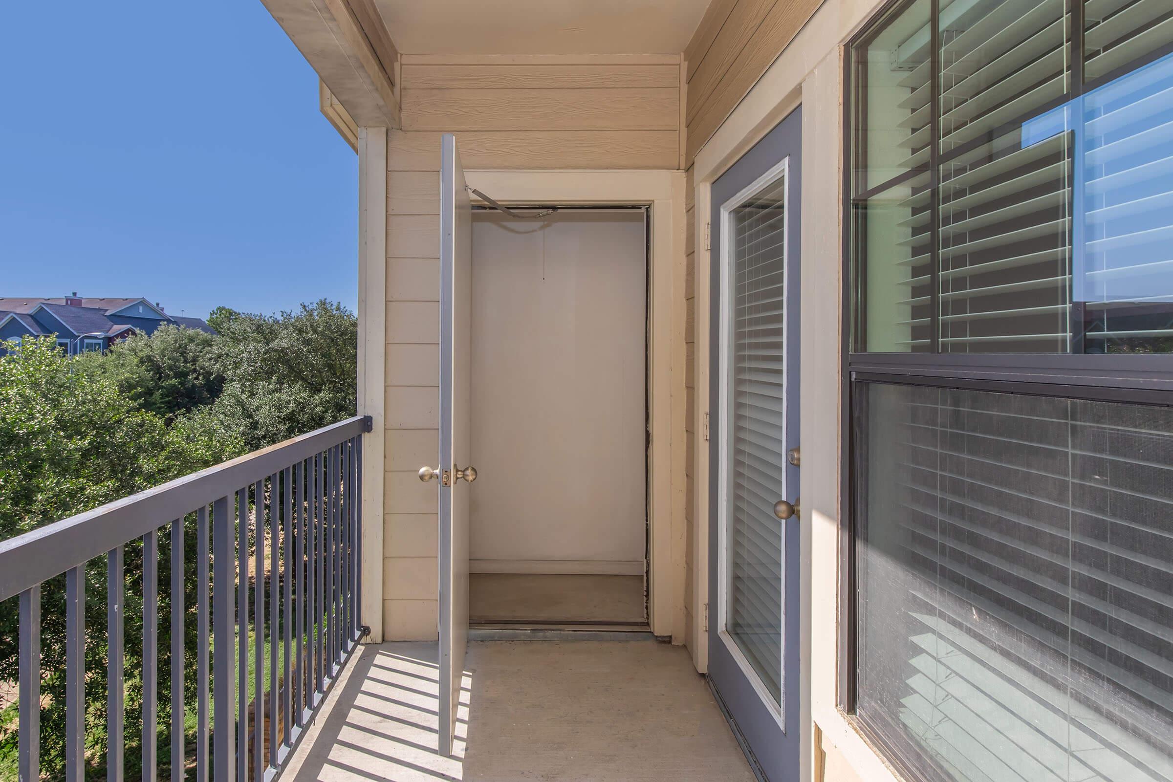A balcony with a railing, featuring two sliding glass doors and a partially open closet door. The backdrop includes green trees and a clear blue sky, suggesting a sunny day. The walls are beige, with horizontal siding, and the floor is concrete, indicating an outdoor space connected to an apartment.