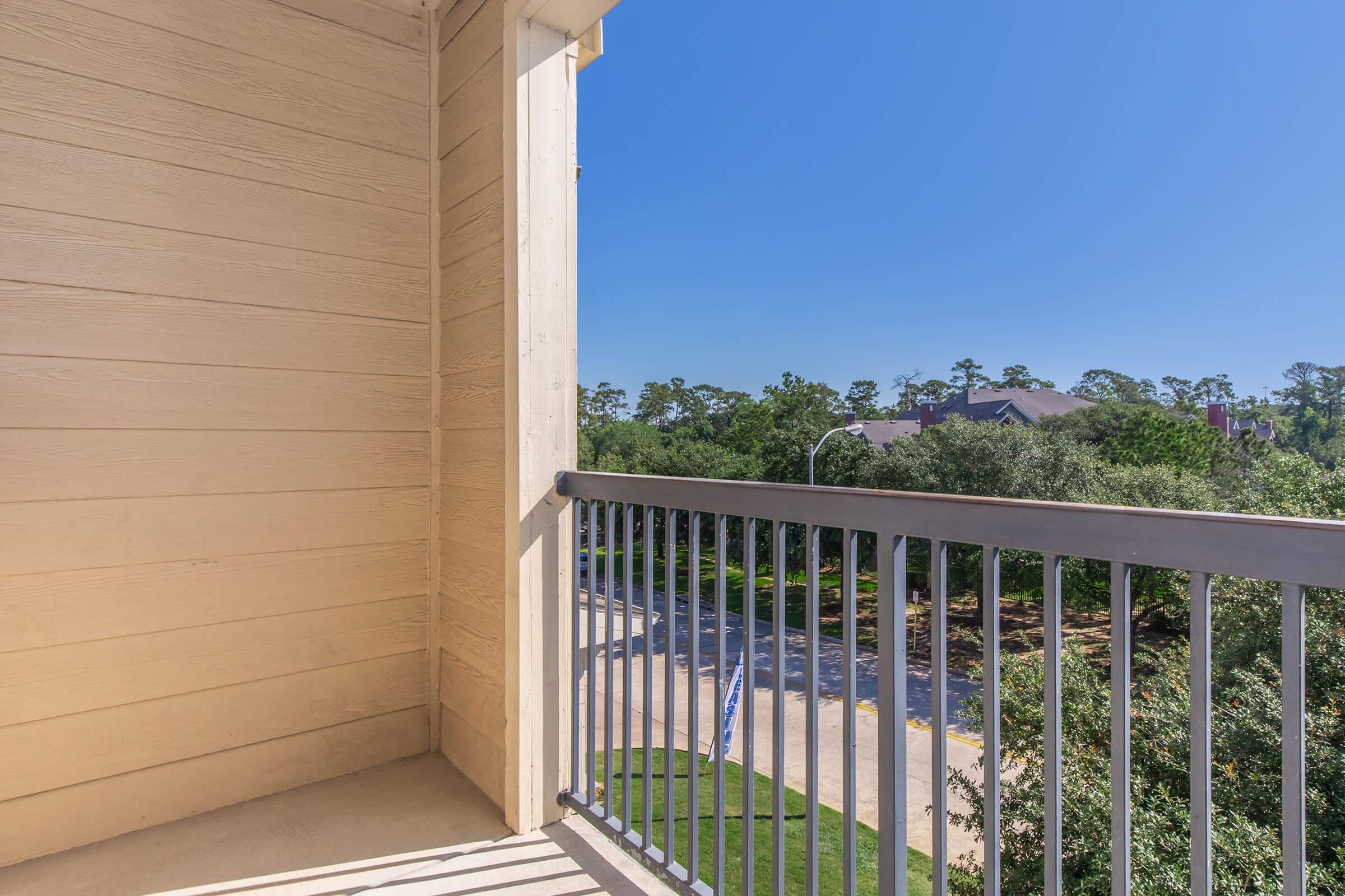 View from a balcony showing a clear blue sky and a tree-lined street below. The balcony is partially enclosed with a railing, and the surrounding area features greenery and residential buildings.