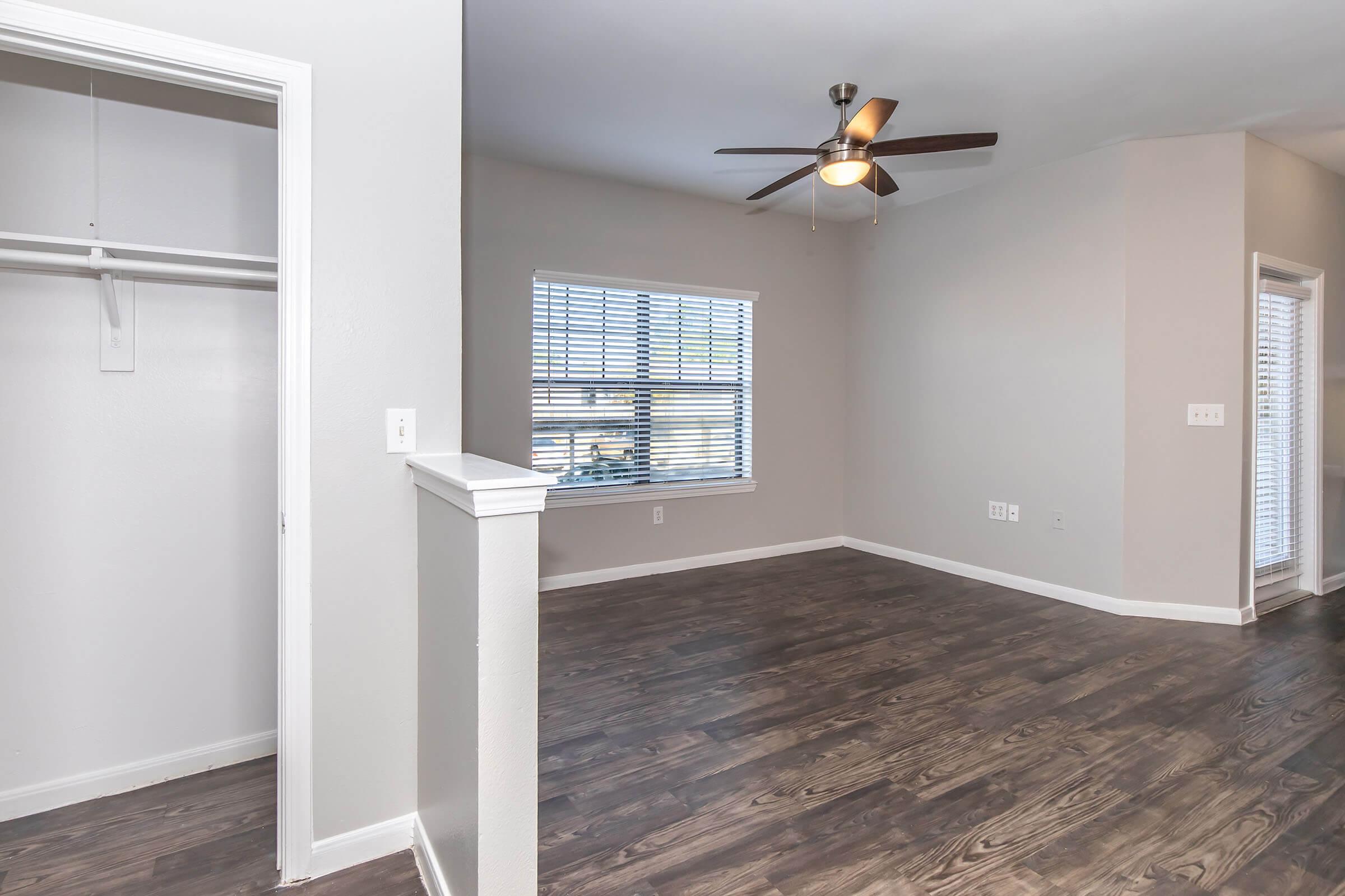 A spacious room with wooden flooring, a ceiling fan, and a window with blinds. A closet is partially visible to the left, and the walls are painted in a light neutral color, creating a bright and airy atmosphere.