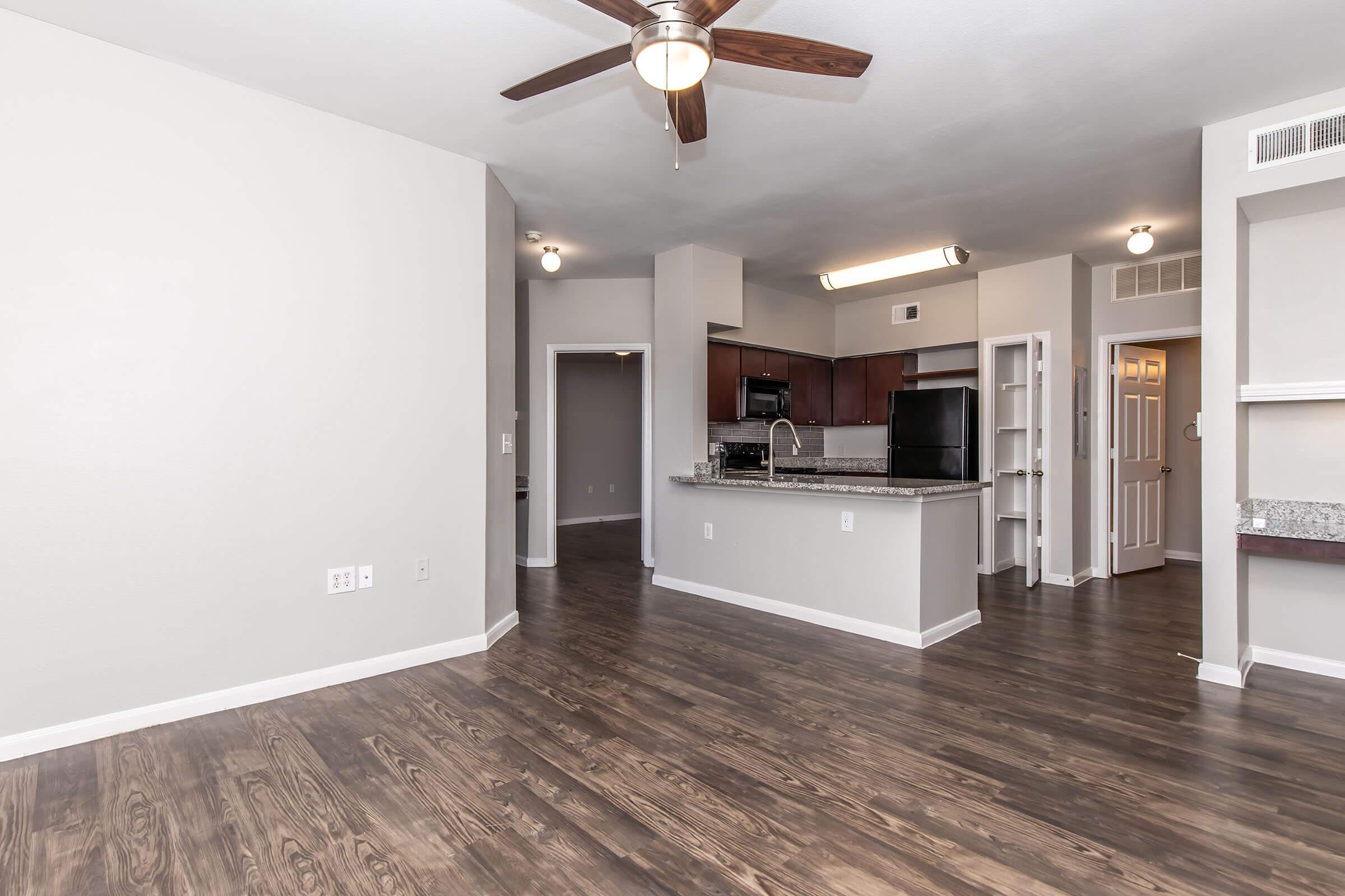Modern apartment interior featuring an open layout with a living area and kitchen. The kitchen is equipped with dark wood cabinets, granite countertops, and stainless steel appliances. The flooring is dark laminate wood. A ceiling fan is present, and there are doorways leading to other rooms in the background.