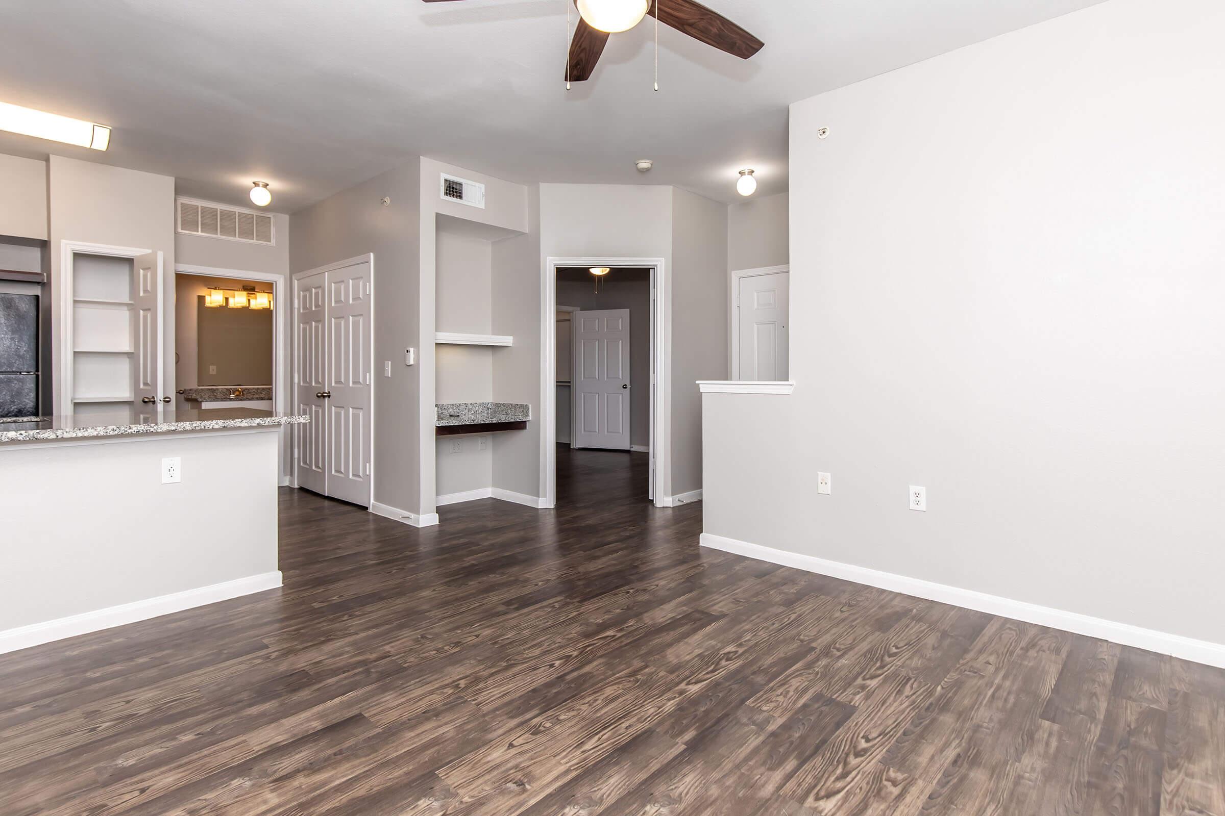 A spacious, modern living area featuring brown hardwood flooring and light gray walls. The room is illuminated with ceiling lights and a ceiling fan. Doorways lead to other rooms, including a bathroom visible in the background. A kitchen area with a breakfast bar is to the left.