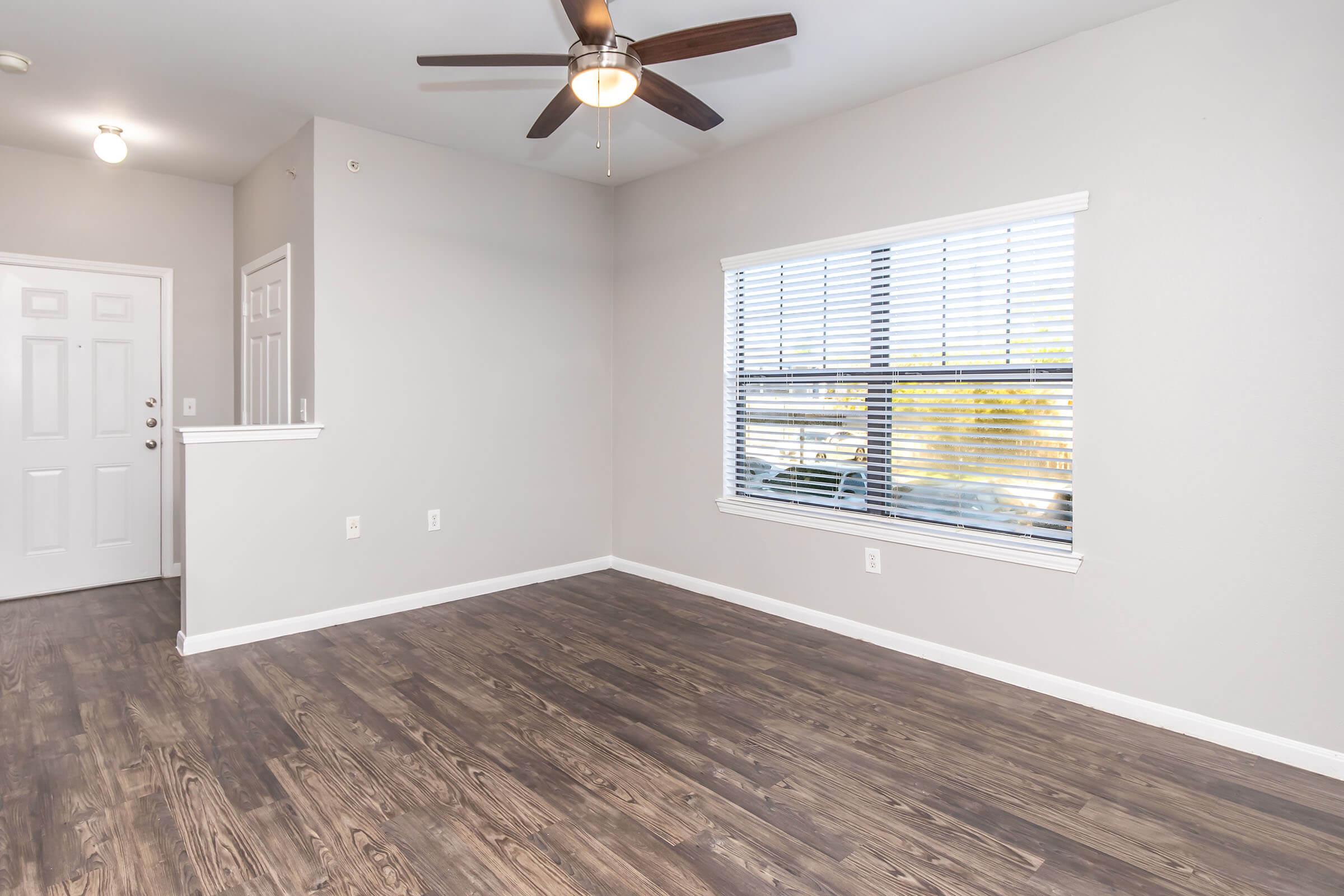 A clean, modern living space with light gray walls and wood-style flooring. There's a ceiling fan on the left side, and a large window with white blinds allowing natural light in. A front door is visible, leading to an entryway. The room appears spacious and empty, ready for furnishing.