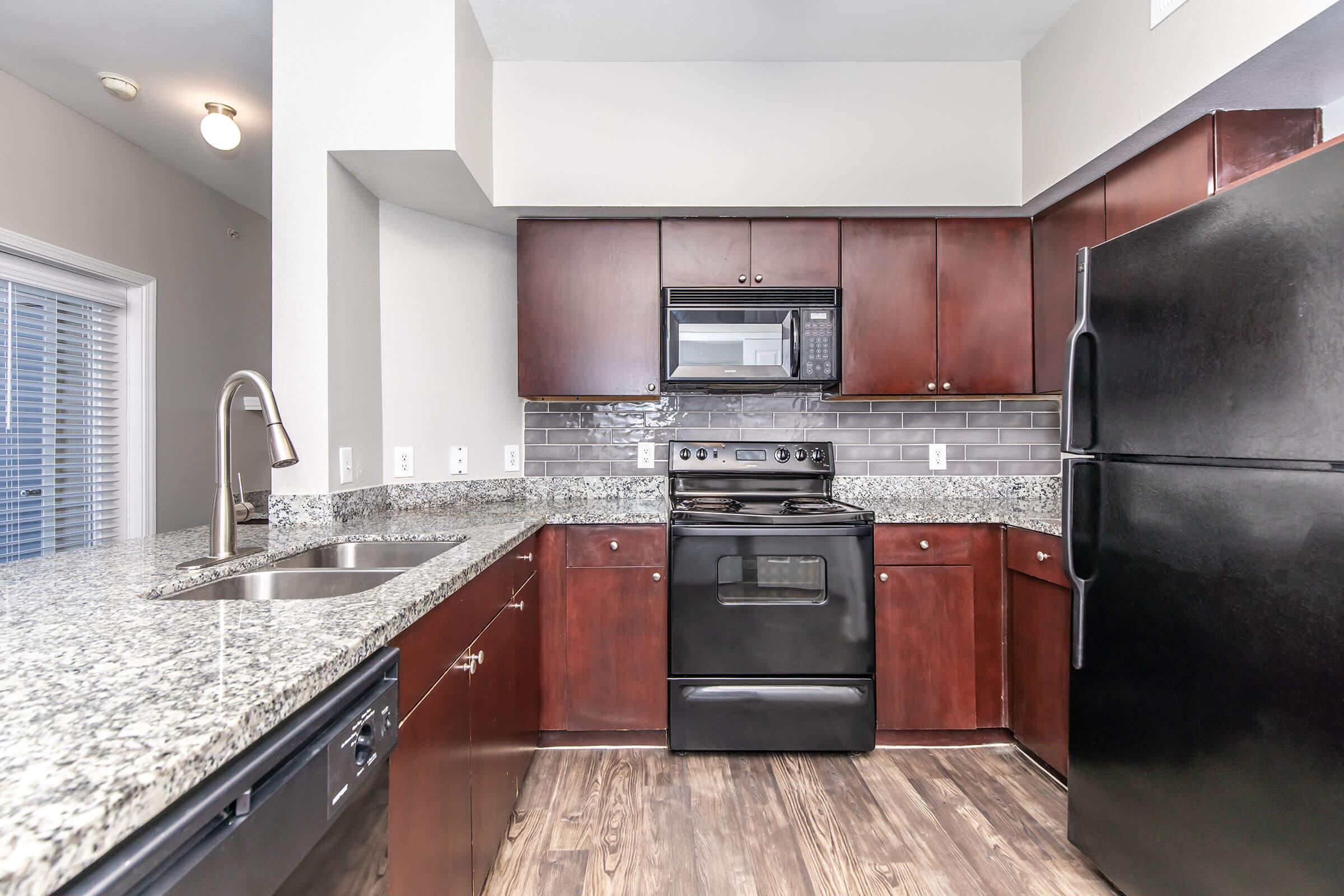 A modern kitchen featuring dark wood cabinets, a black refrigerator, and an oven with a microwave above. The granite countertop adds a sleek touch, and the backsplash consists of gray tiles. Natural light comes in through a window, enhancing the open-space feel. The flooring is a warm wood finish.