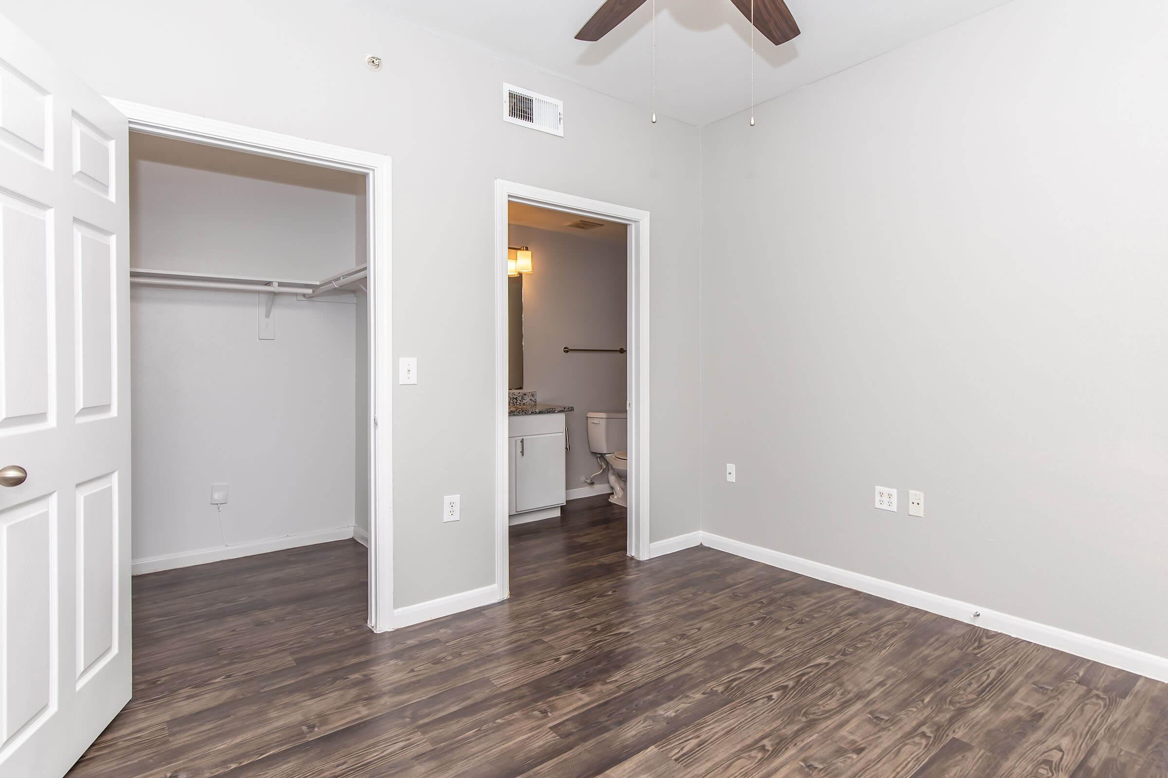 An empty room featuring gray walls and dark wood flooring. There is a ceiling fan and two doorways: one leading to a closet and the other to a bathroom. The overall ambiance is bright and minimalistic.