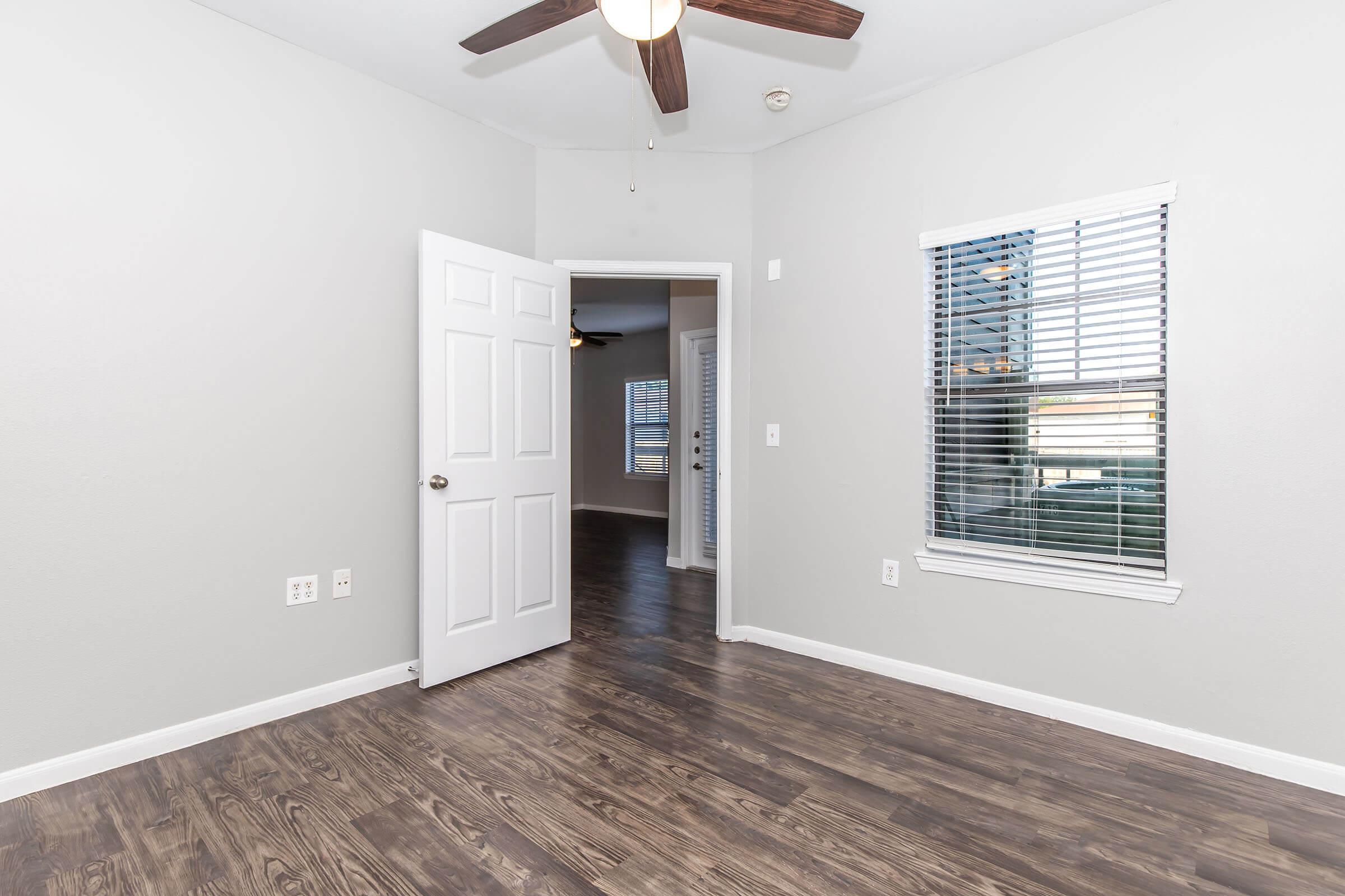 A well-lit interior of a room with light gray walls and a dark wood floor. There is a ceiling fan above, a white door partially open leading to another room, and a window with blinds allowing natural light. The space appears neat and organized, suitable for living or workspace.