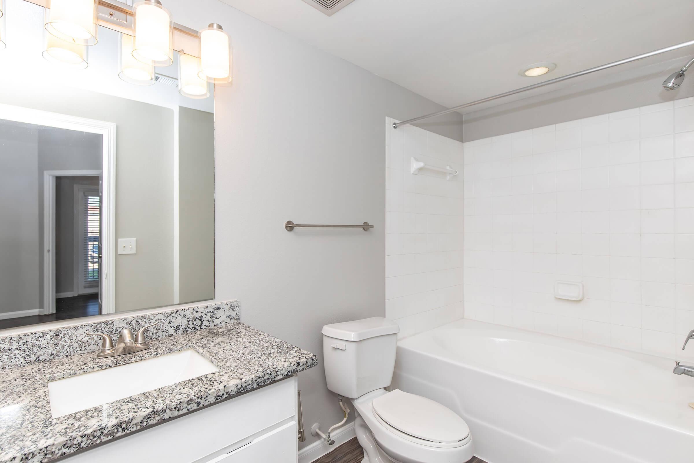 A modern bathroom featuring a white bathtub with a shower, a sleek granite countertop with a sink, a toilet, and a lit mirror. The walls are painted light gray, and there is a towel bar mounted on the wall. The space is well-lit and clean, with a view into another room beyond the bathroom.