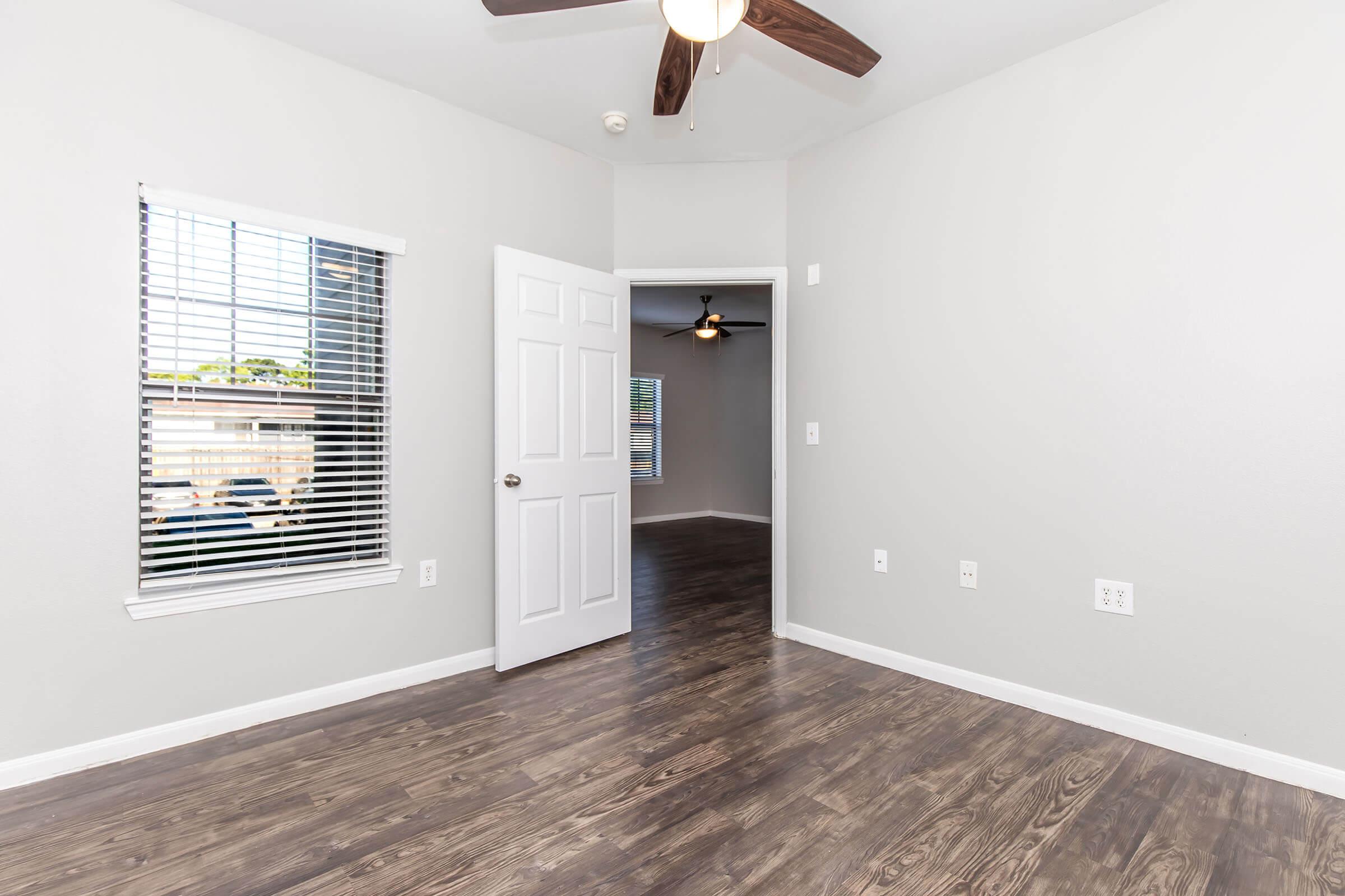 Interior of a light-colored room featuring a ceiling fan, wooden flooring, and large windows with blinds. A doorway leads to another room, and the walls are painted in a neutral tone. The space appears bright and inviting, ideal for various uses.
