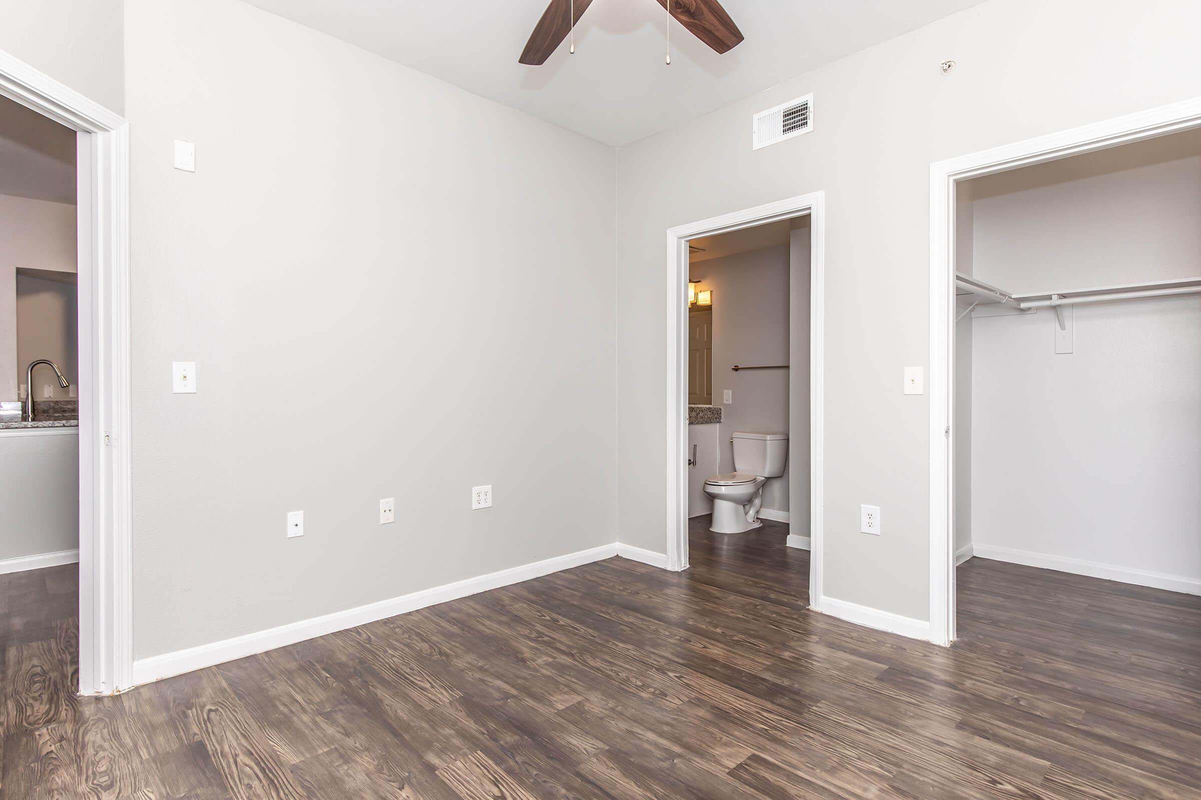 A spacious, unfurnished room with light gray walls, featuring a ceiling fan and a wood-style floor. Two doorways lead to a bathroom and a closet, enhancing the room's functionality. Natural light may be present, creating an inviting atmosphere.