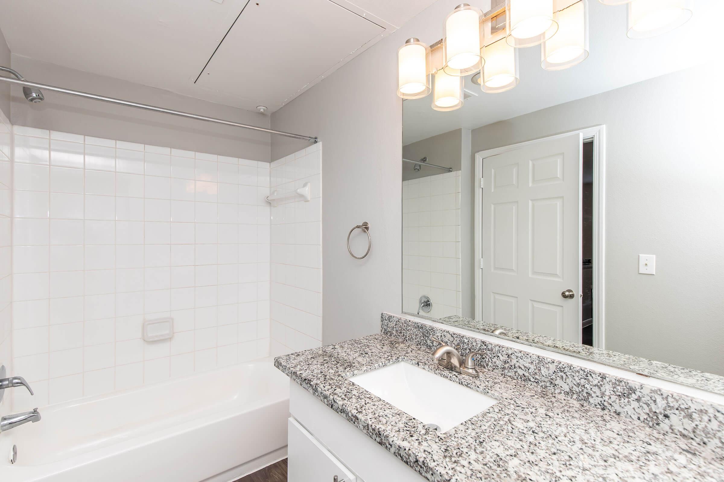 Modern bathroom featuring a white tiled bathtub with a shower, a granite countertop sink with a faucet, and a wall mirror. Illuminated by a stylish light fixture above. The walls are painted in a light gray, and there's a white door visible in the background.