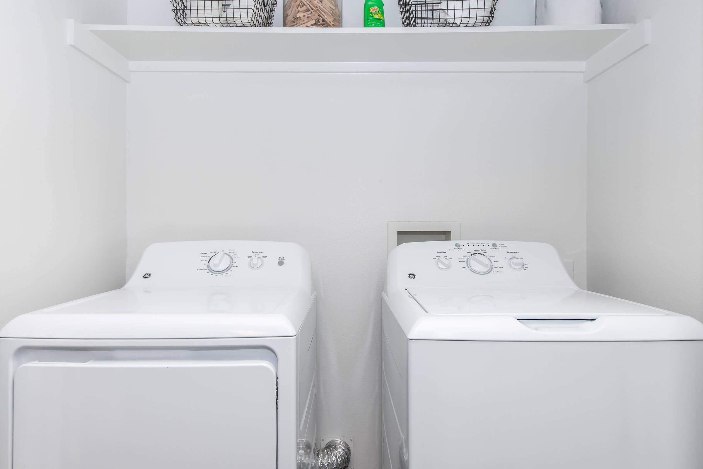 A laundry room featuring a front-loading washing machine next to a top-loading dryer. Above them, a shelf holds wire baskets and a laundry detergent container. The walls are painted white, and the appliances are neatly arranged, creating a clean and organized space.