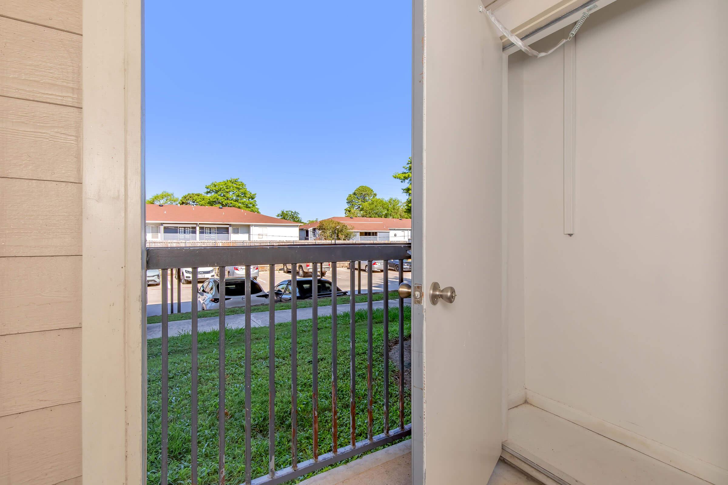 View from an apartment balcony featuring an open door, a railing, and a glimpse of the surrounding green lawn and parked cars below. The sky is clear and blue, with buildings visible in the background. The space appears bright and inviting.