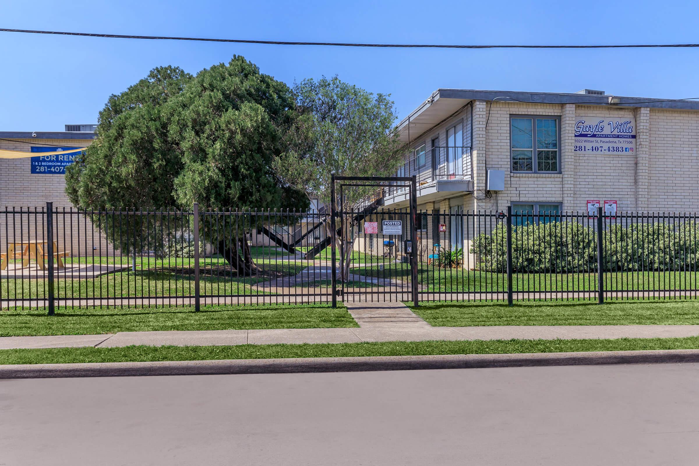A gated exterior view of a two-story building surrounded by greenery. A large tree is visible in the foreground, with a fence enclosing the area. Signs on the building indicate rental information. The scene is bright and sunny, with a clear blue sky.