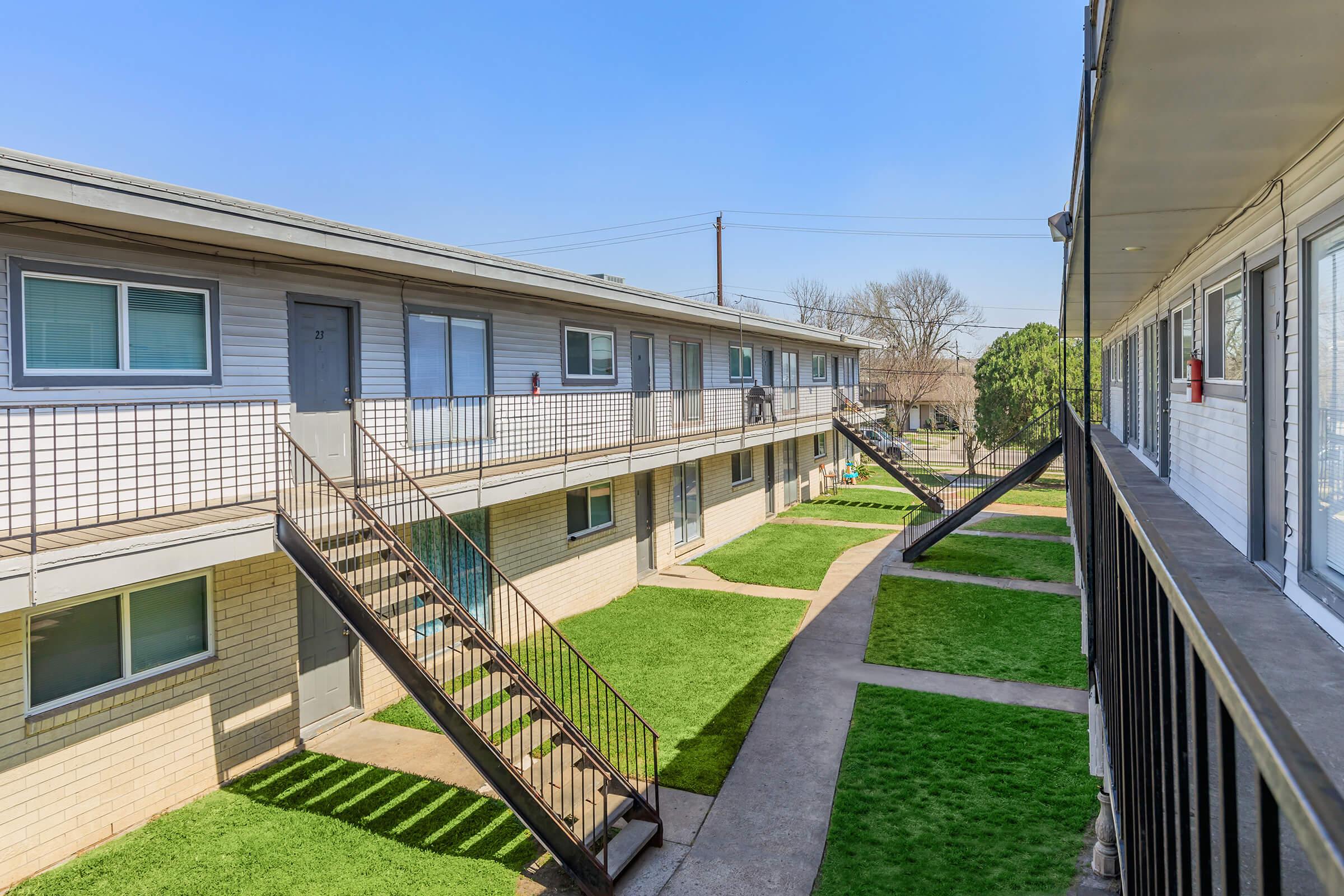 View of a multi-unit apartment complex featuring two-story buildings arranged in a U-shape. Stairs lead to second-floor units, with green lawns and concrete pathways between them. Clear blue sky above, indicating a sunny day.