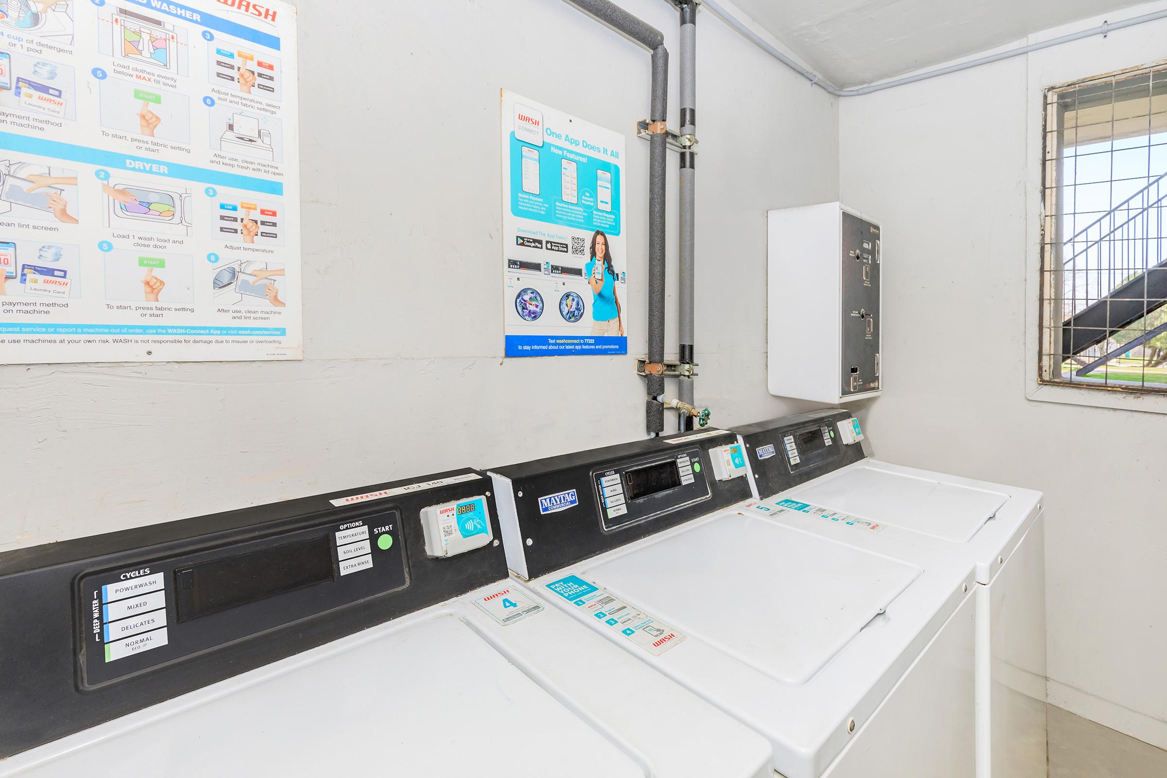 A laundry room featuring multiple washing machines and a dryer. There is a wall-mounted instructional poster explaining machine use. The room is well-lit with a window, and the machines are modern, displaying various buttons and settings for different laundry options.