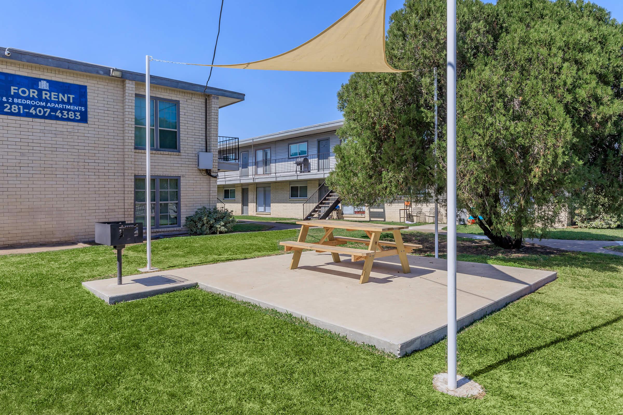 A picnic table with an umbrella is situated on a concrete pad in a green grassy area. In the background, a two-story apartment building is visible with a "For Rent" sign. There are steps leading to the upper floor, and another building is seen to the right. Clear blue sky overhead.