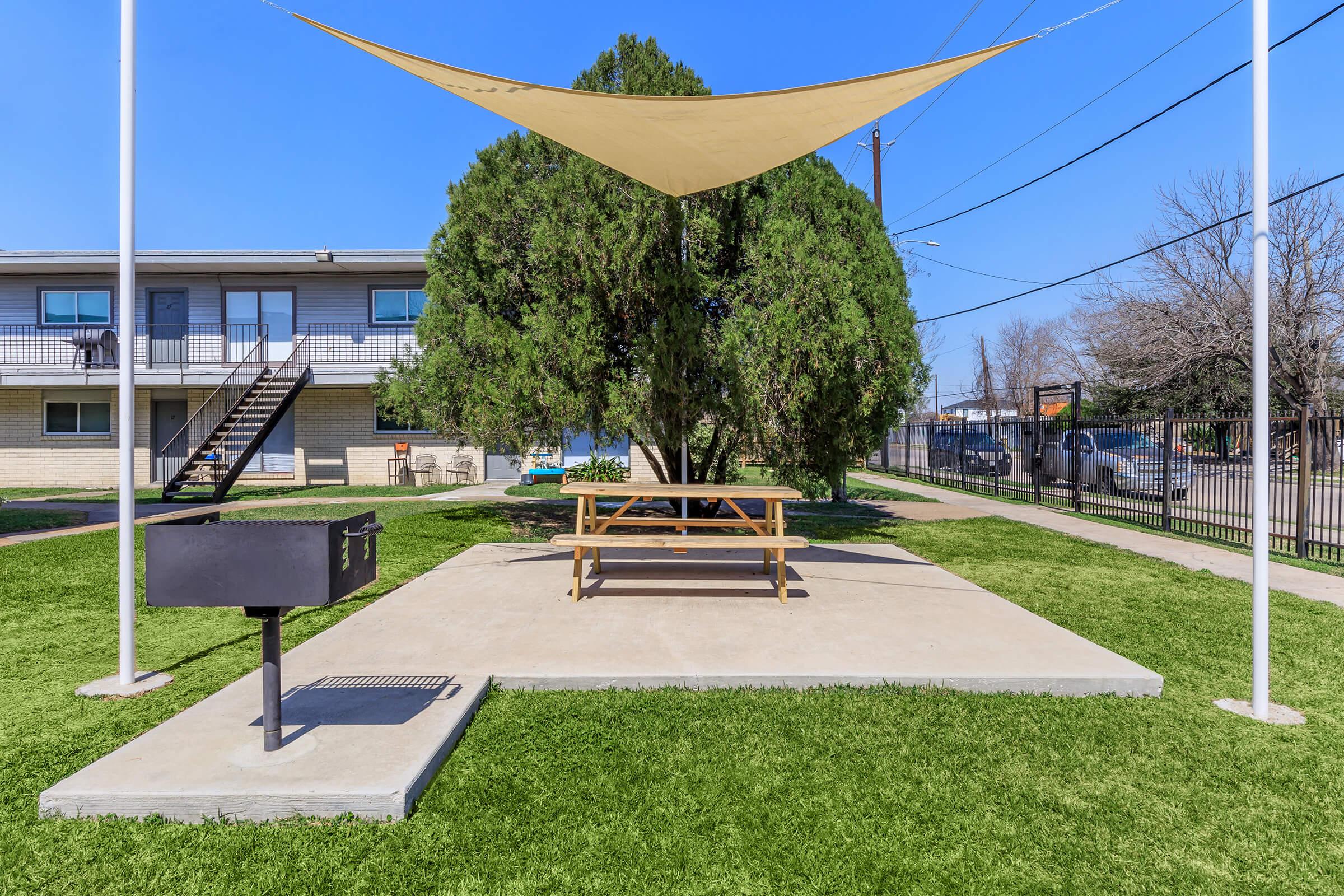 A picnic area featuring a concrete table and benches, shaded by a large fabric canopy. A charcoal grill is positioned next to the table, surrounded by green grass. In the background, there is a multi-unit building and utility poles under a clear blue sky.
