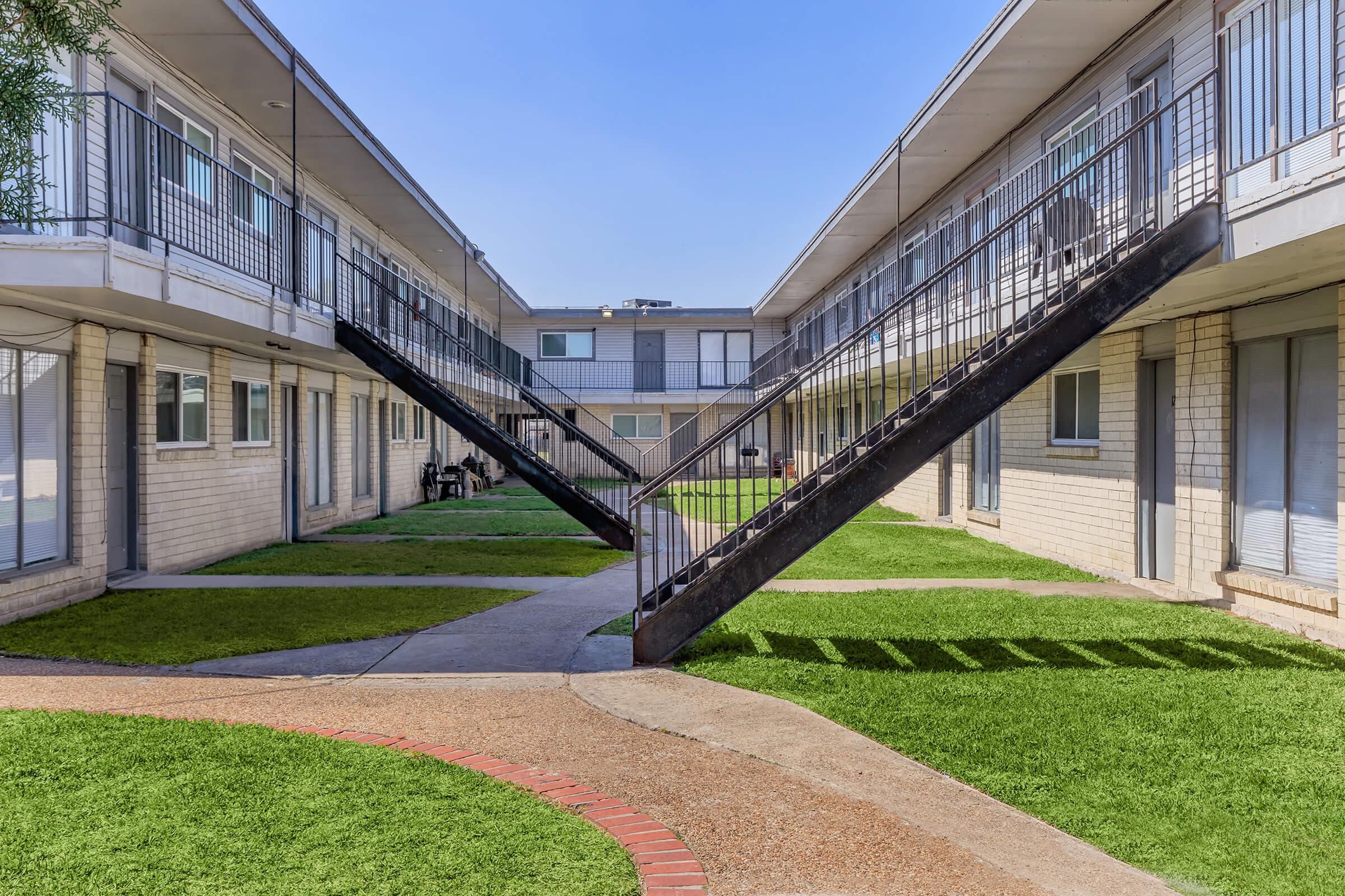 A view of a two-story apartment complex featuring a central courtyard with green grass. The buildings have staircases leading to the upper levels, and the walkway is lined with shadows cast by the railings. Clear blue sky overhead enhances the scene.