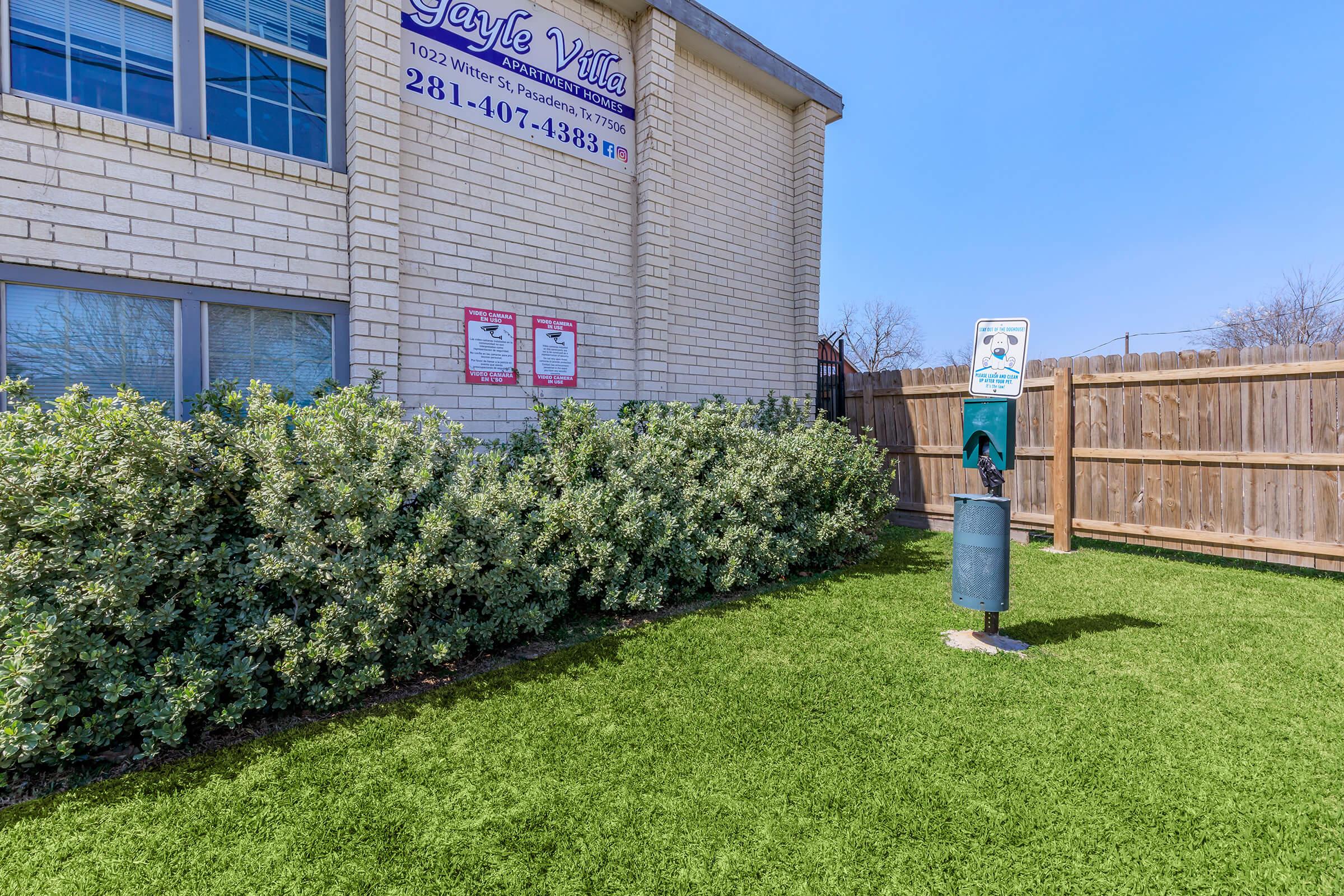 A well-maintained outdoor area featuring artificial grass, a bushy shrub, and a pet waste disposal station next to a building with signage. The fence provides privacy to the space, enhancing the overall cleanliness and organization of the environment.