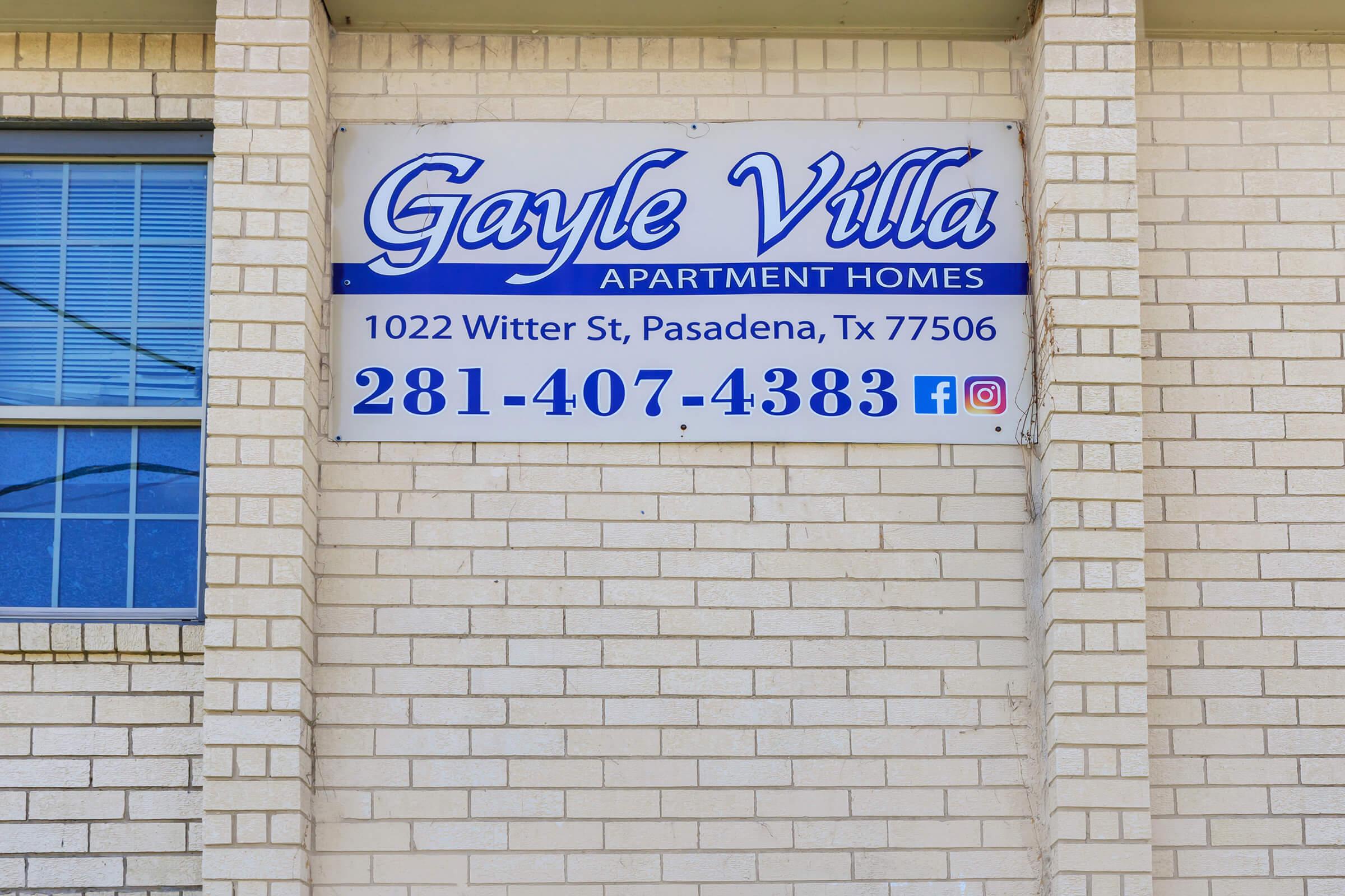 Sign for Gayle Villa Apartment Homes located at 1022 Witter St, Pasadena, TX 77506, featuring the phone number 281-407-4383 and social media icons. The sign has a blue and white color scheme and is mounted on a brick wall with windows above.