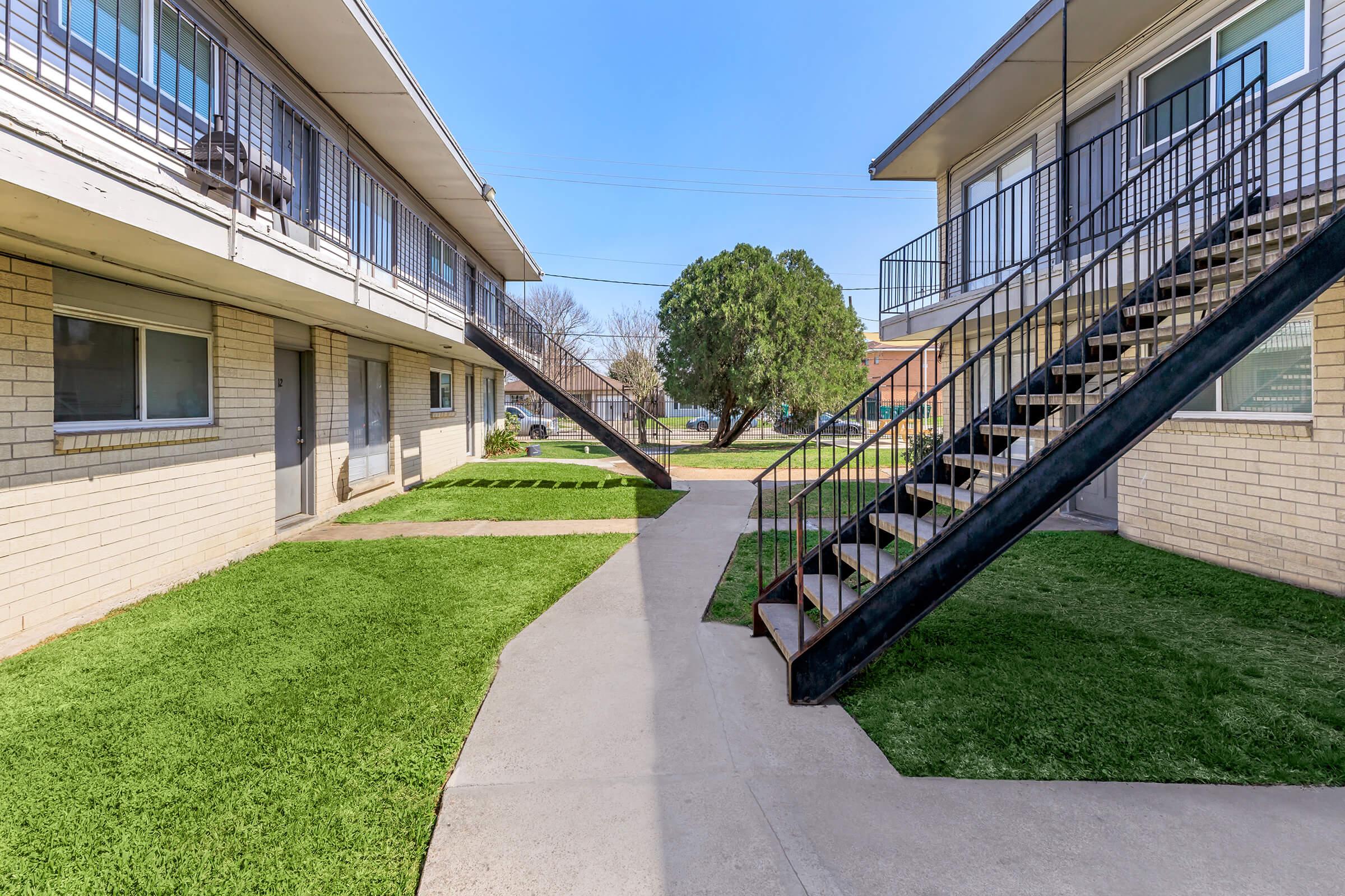 A view of a residential courtyard featuring two sets of stairs leading to upper-level apartments. The area is landscaped with green grass and concrete walkways. The building exterior is brick, and the sky is clear and bright. Trees are visible in the background, adding to the inviting atmosphere.