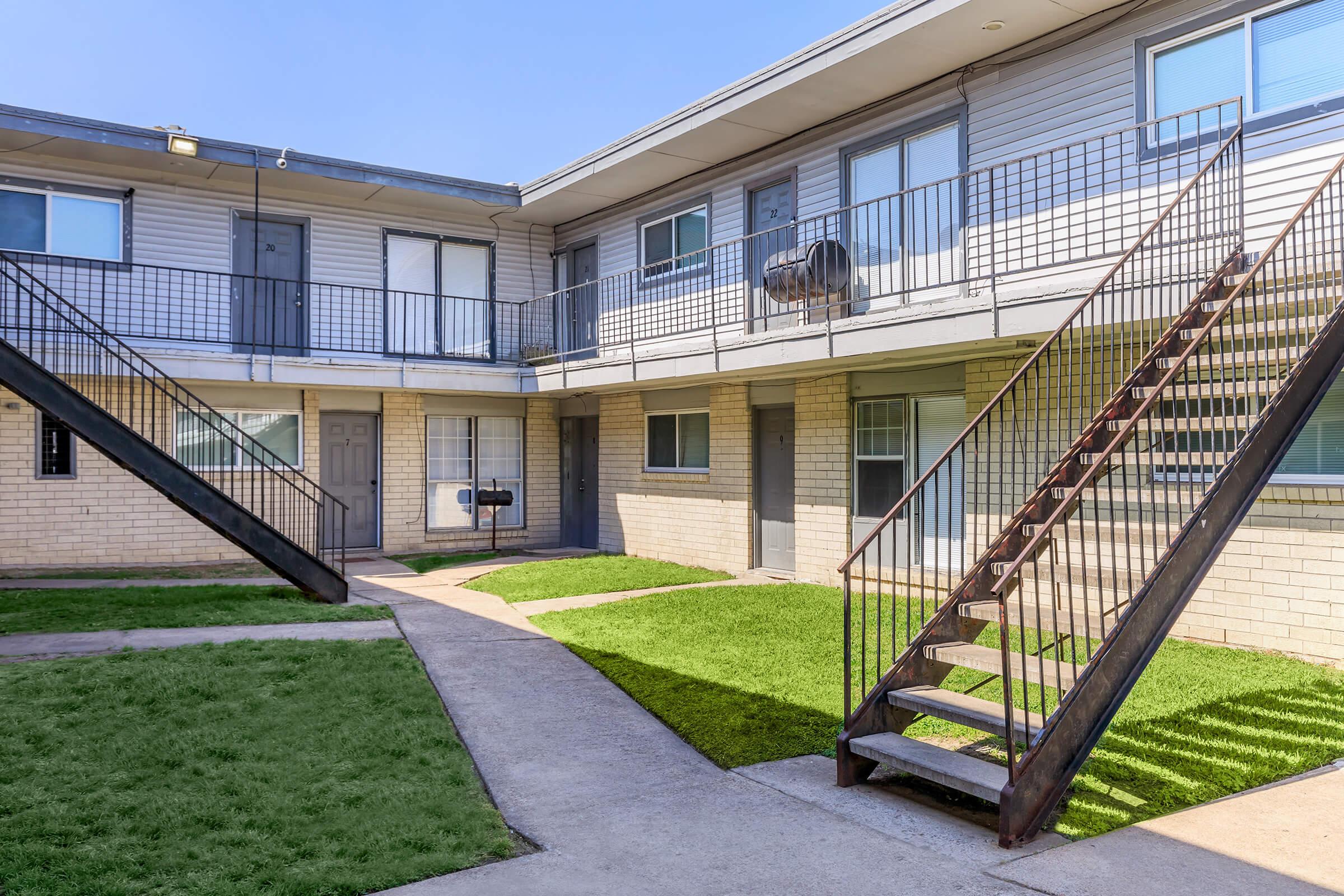 A view of an apartment complex courtyard featuring two sets of metal stairs leading to upper-level units. The ground is covered with green grass, and there are small pathways leading to doors of the apartments. The building has a modern design with multiple windows and air conditioning units.