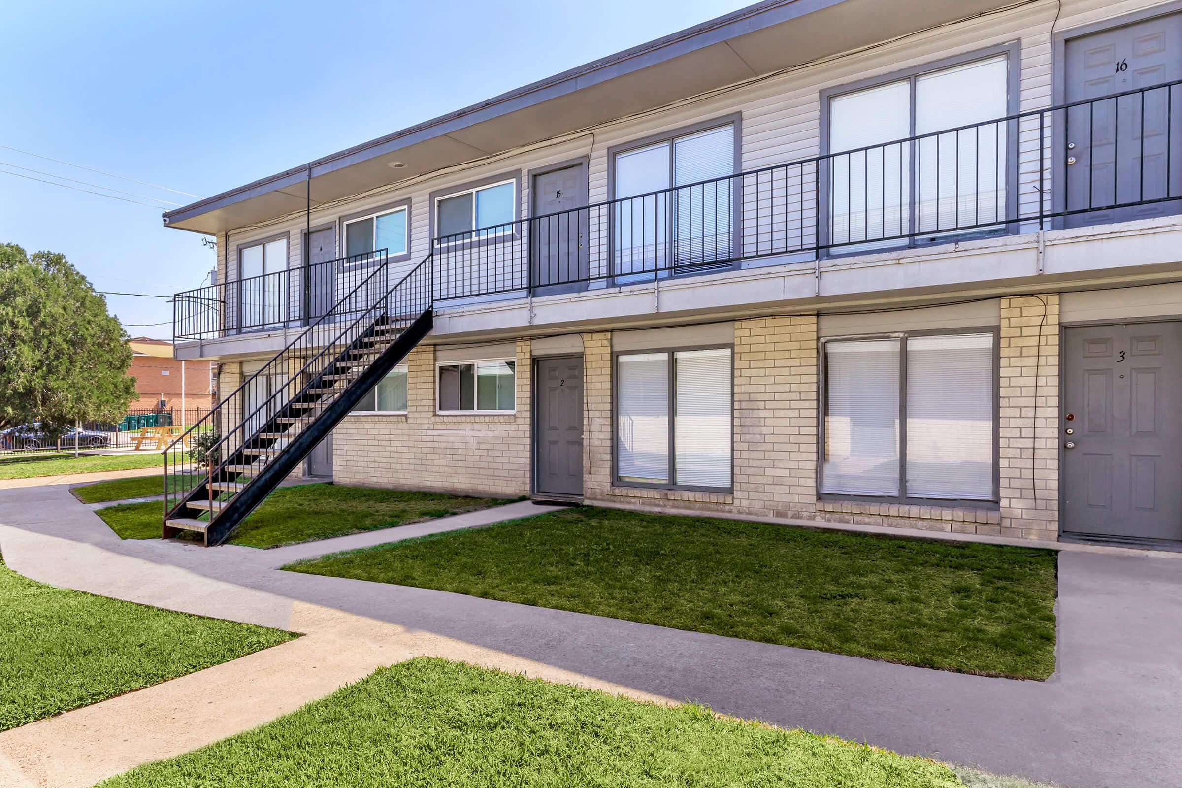 A two-story apartment building featuring multiple units with grey doors and windows. A stairway leads to the second floor, and a well-maintained pathway runs alongside a grassy area in front of the building. The setting is bright with clear blue skies.