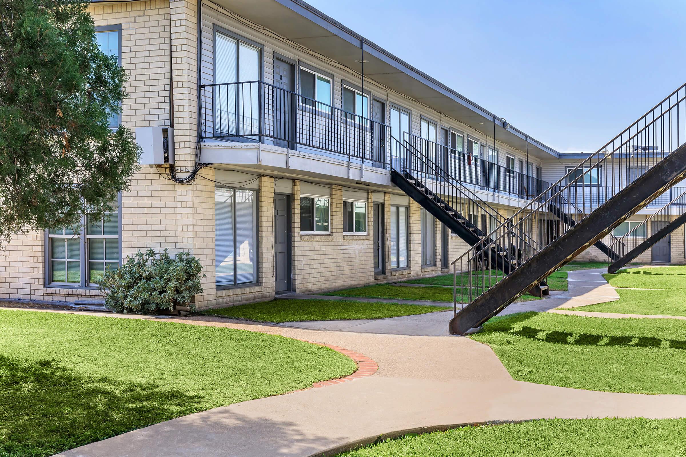 A view of a two-story apartment building with a central grassy courtyard. The building features several windows and balconies, with a black metal staircase leading to the upper floor. There is a walkway made of concrete leading through the green lawn. The sky is clear and blue.