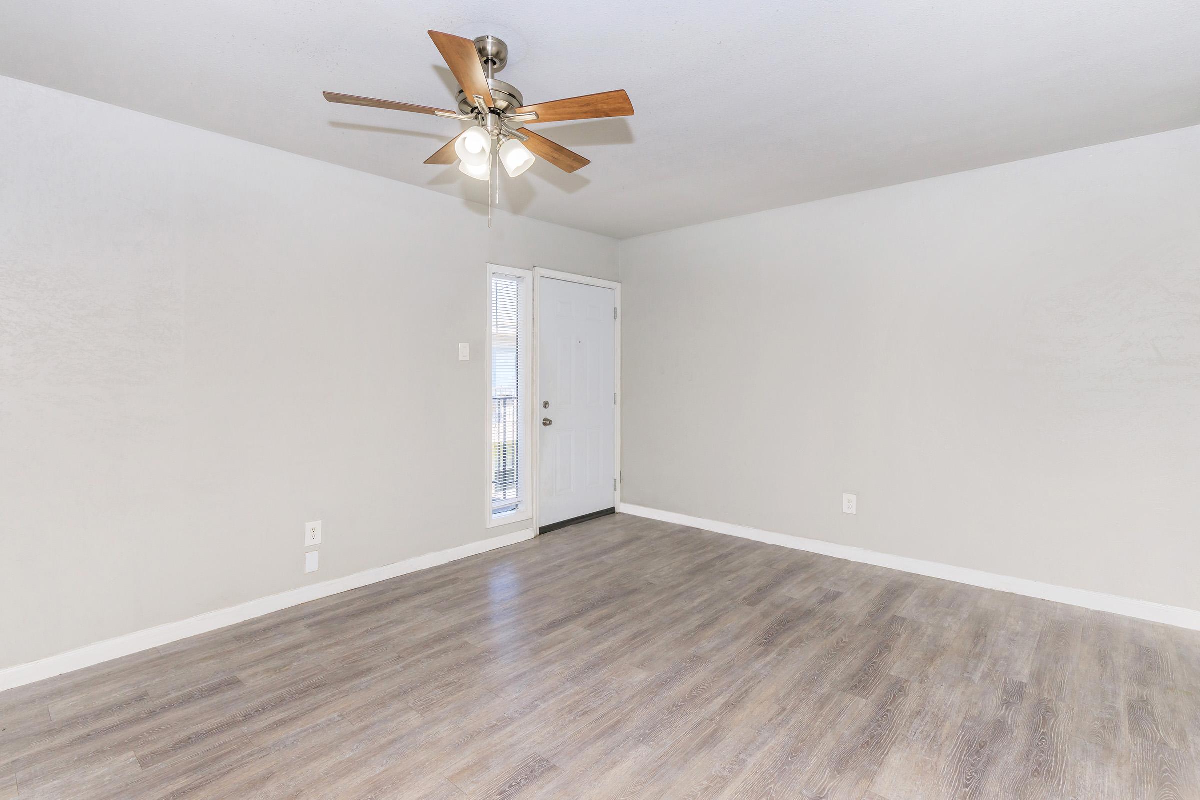 A spacious room with light gray walls and a light wooden floor. It features a ceiling fan with wooden blades and a white light fixture. A door is visible on the left, leading outside, while a window provides natural light. The overall ambiance is clean and inviting, suitable for various furnishings.