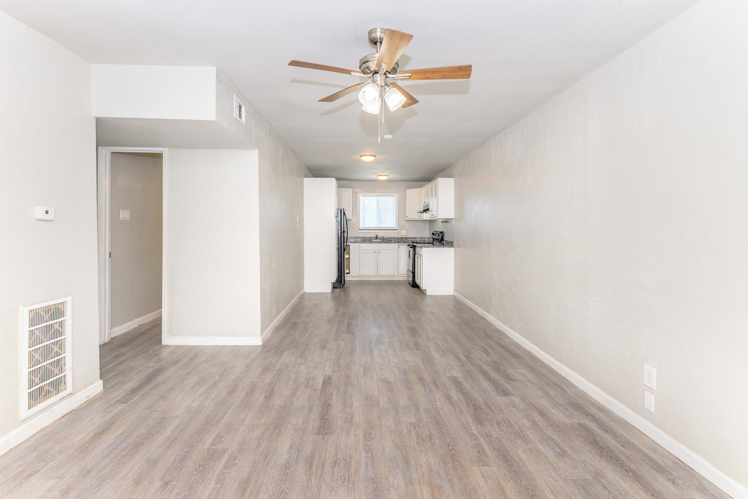 A spacious, modern living area with light-colored walls and a wood-patterned floor. A ceiling fan hangs above, and to the right, a white kitchen is visible, featuring a refrigerator and sink. The room is well-lit, with a large window allowing natural light, creating an open and airy atmosphere.