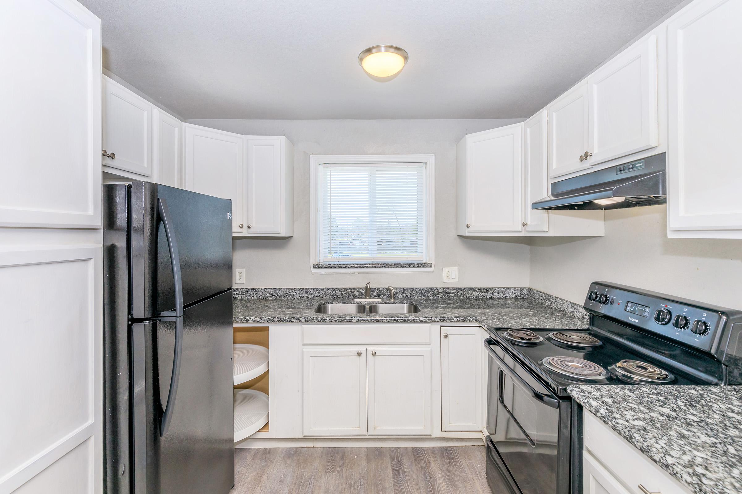 Modern kitchen featuring white cabinetry, a black refrigerator, and a black stove with an oven. The countertops are speckled gray, and there's a window allowing natural light. The space is tidy and functional, with a sink between the stove and the window.