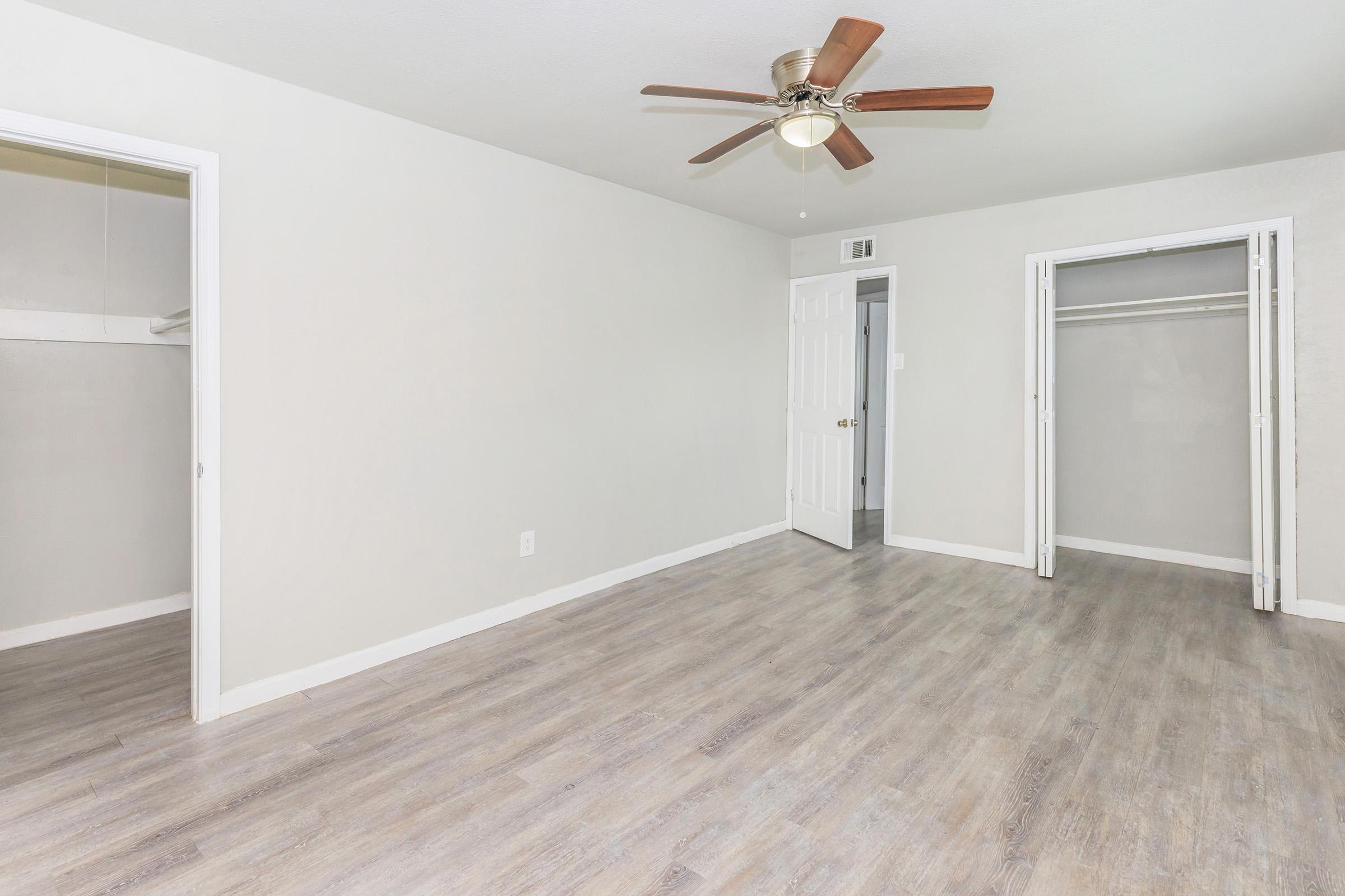 A spacious, minimally furnished room with light gray walls and a ceiling fan. The room has light wooden flooring, a closed door leading to a bathroom, and two open closets on the right side. Natural light fills the space, making it bright and inviting.