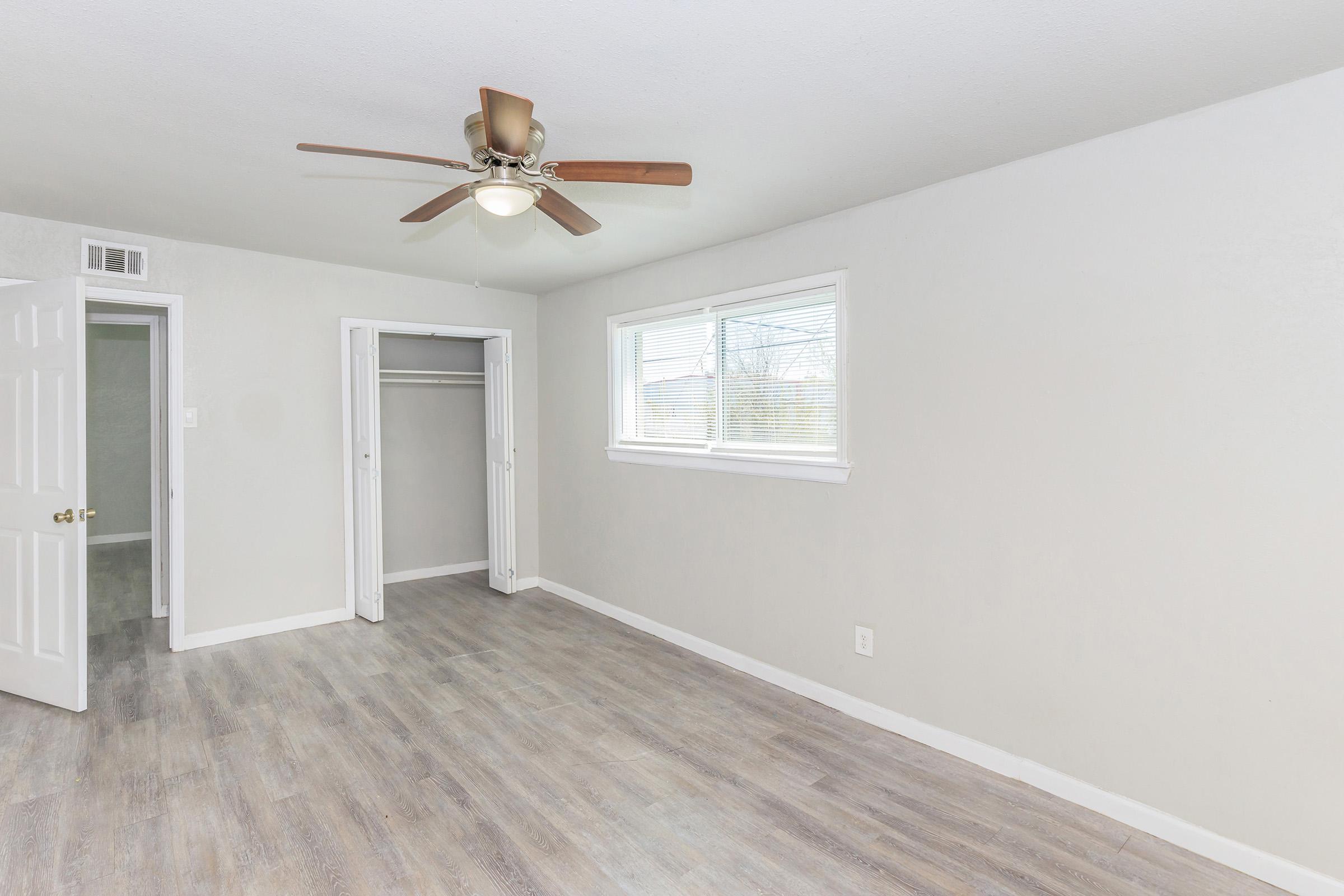 A bright, empty room featuring light gray walls, a ceiling fan, and a light wood floor. There is a window allowing natural light, and an open closet with double doors on one side. The space is clean and ready for furniture or decoration.