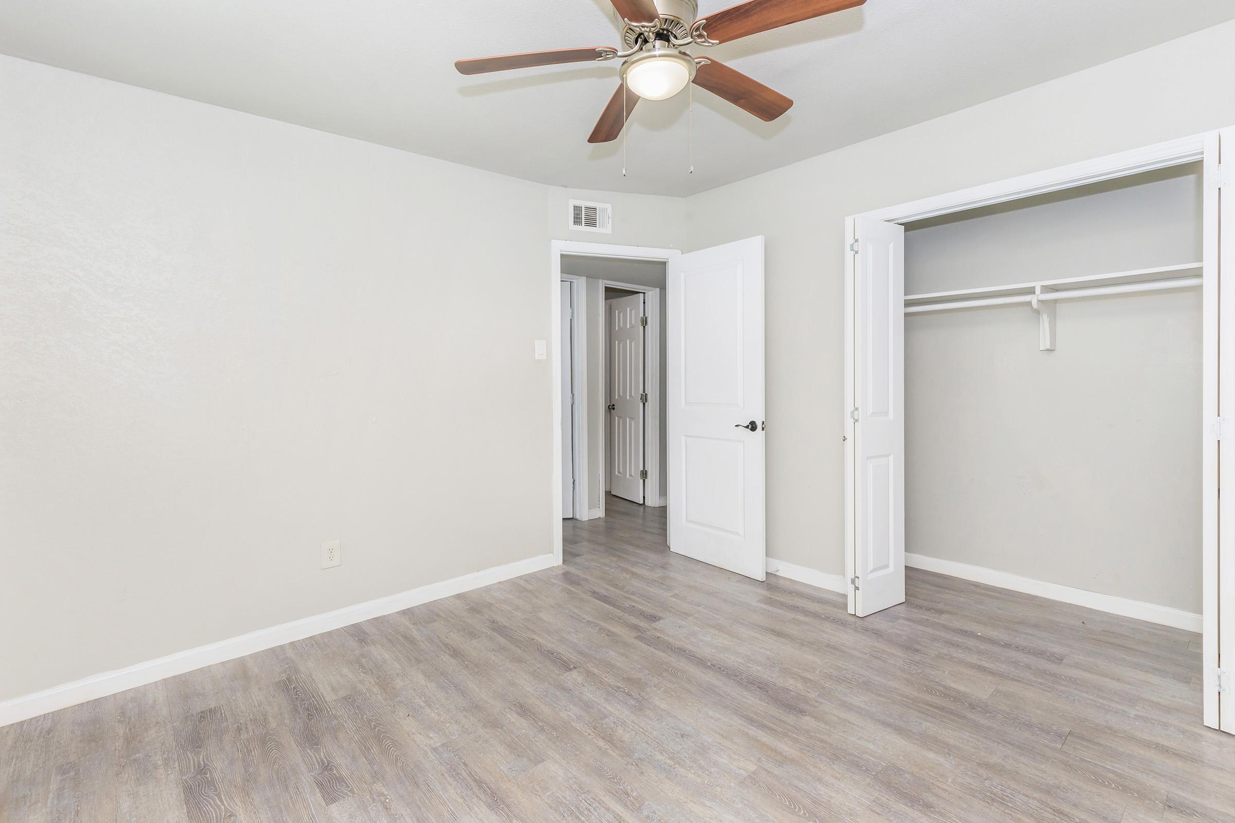 Spacious, empty room with light gray walls and a ceiling fan. The flooring is light wood laminate. There’s a closet with open double doors on the left, and a door leading to another room in the background. The overall ambiance is bright and inviting, ideal for personal decoration.