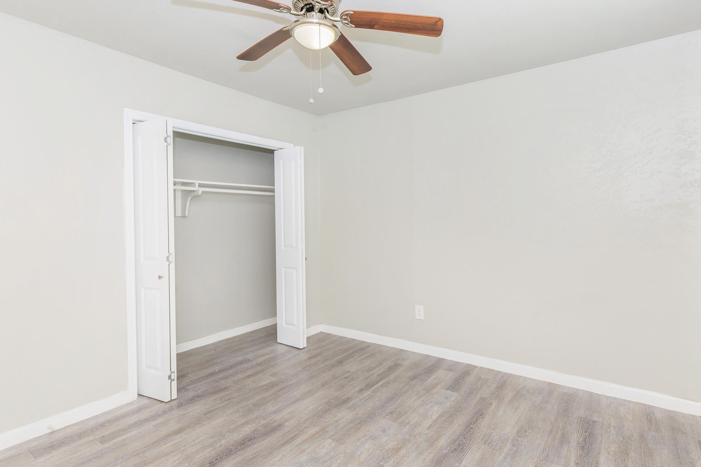 Interior of a minimalist room featuring light gray walls, a ceiling fan with wooden blades, and light wood flooring. The room includes a closet with double doors on the left side, and ample open space, creating a bright and airy atmosphere.
