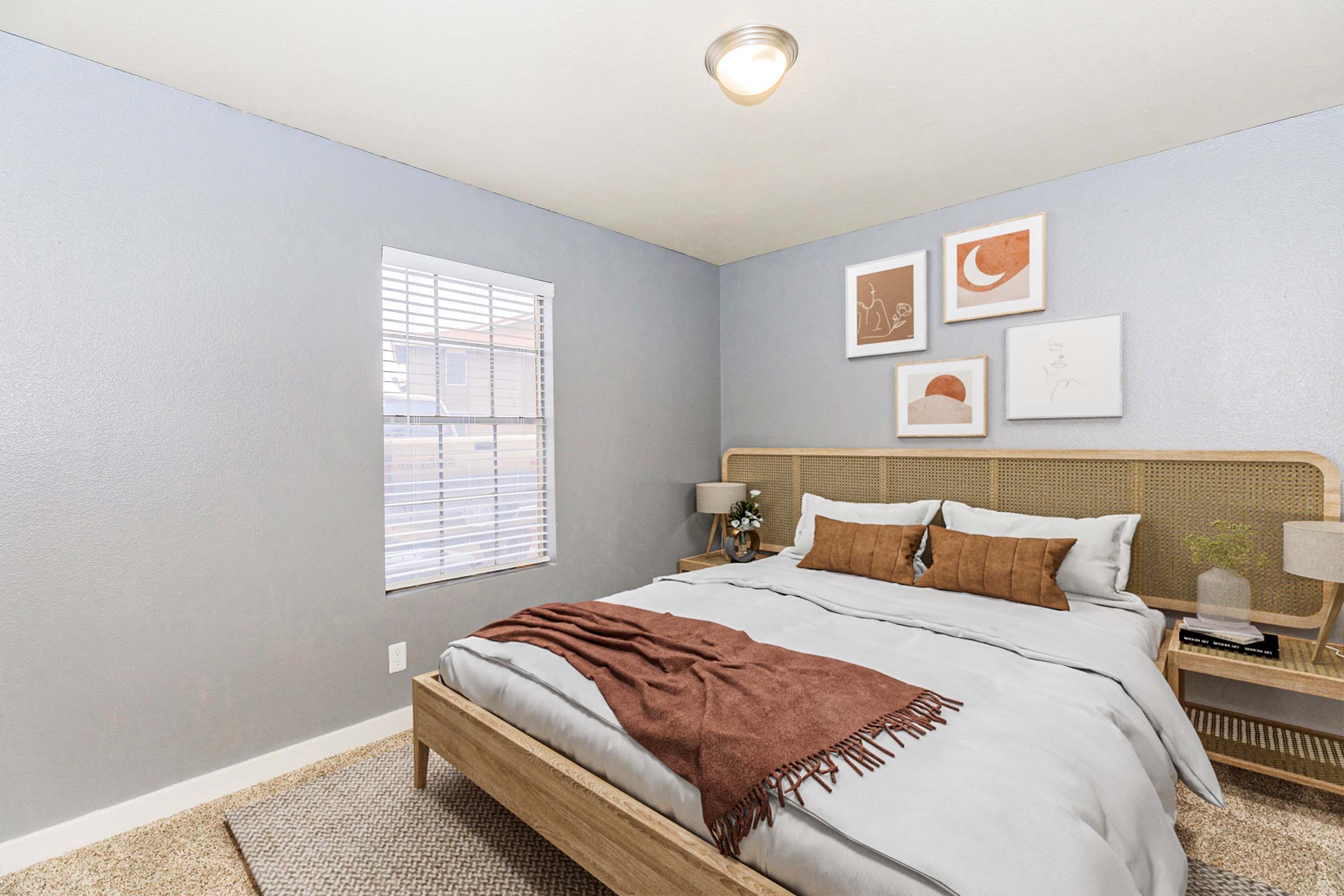 A cozy bedroom featuring a neatly made bed with gray bedding and decorative pillows. There are two small bedside tables with lamps, and a window letting in natural light. The walls are painted a soft gray, and above the bed are three framed artworks in neutral tones. A beige rug covers the floor.