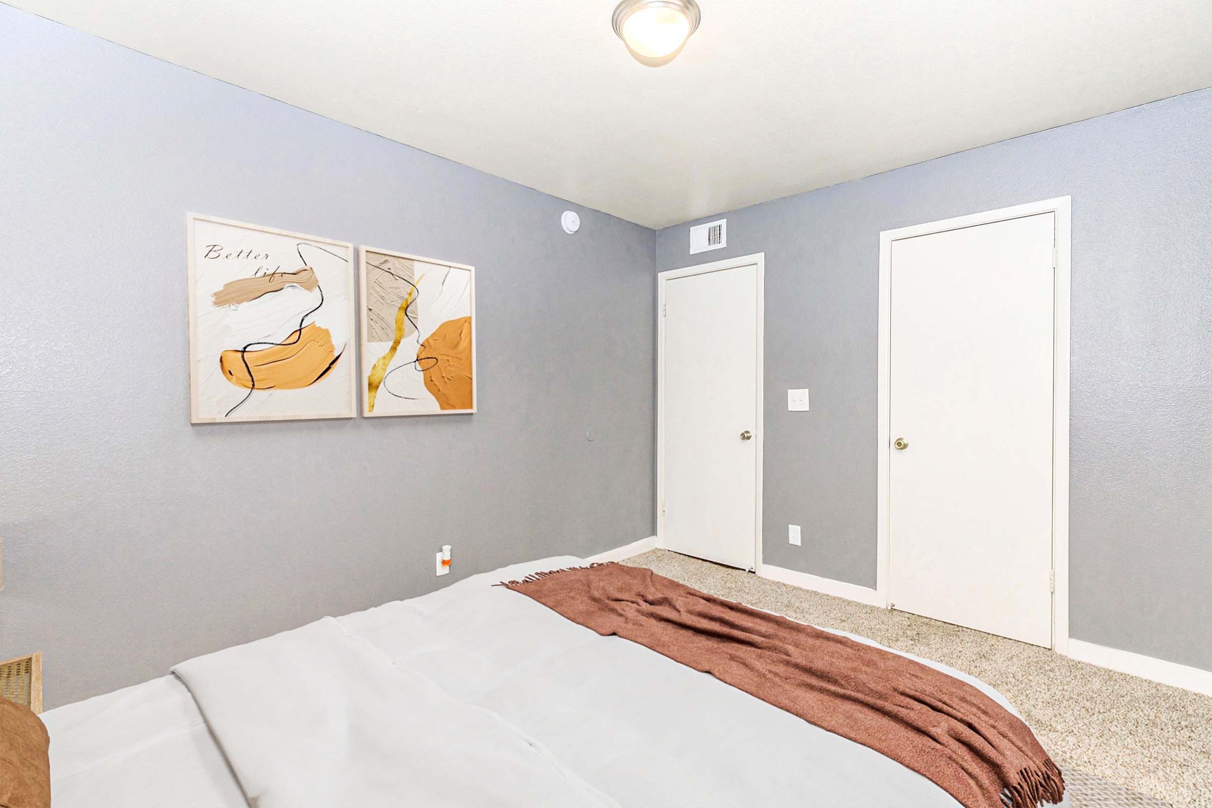 A cozy bedroom featuring a neatly made bed with a light gray blanket and a brown throw. Two abstract art pieces hang on the wall, complementing the soft gray walls. Two doors are visible, and the room has carpet flooring, providing a warm atmosphere. A ceiling light adds brightness to the space.